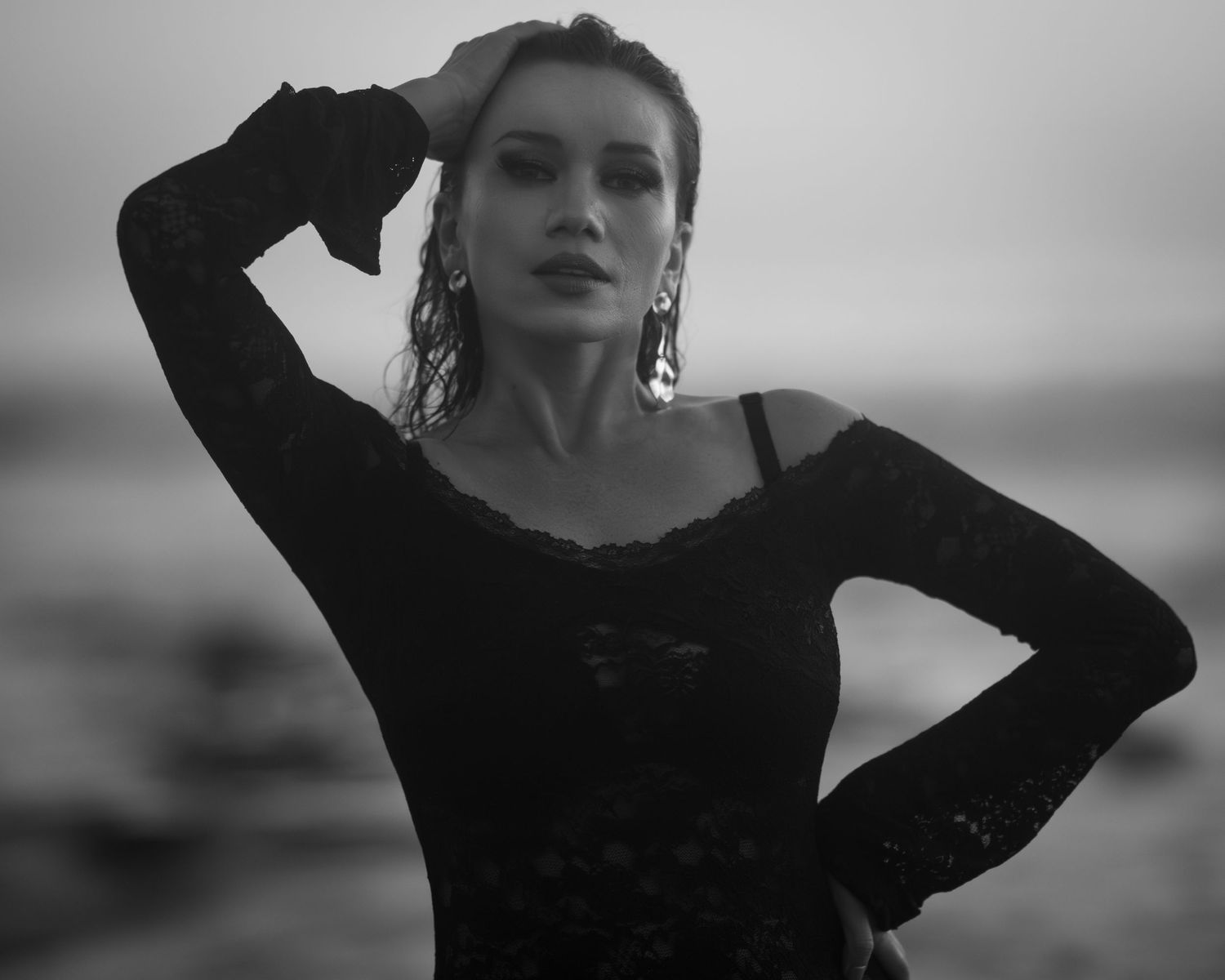 Black and white photo of an elegant woman in lace dress posing dramatically on a beach.