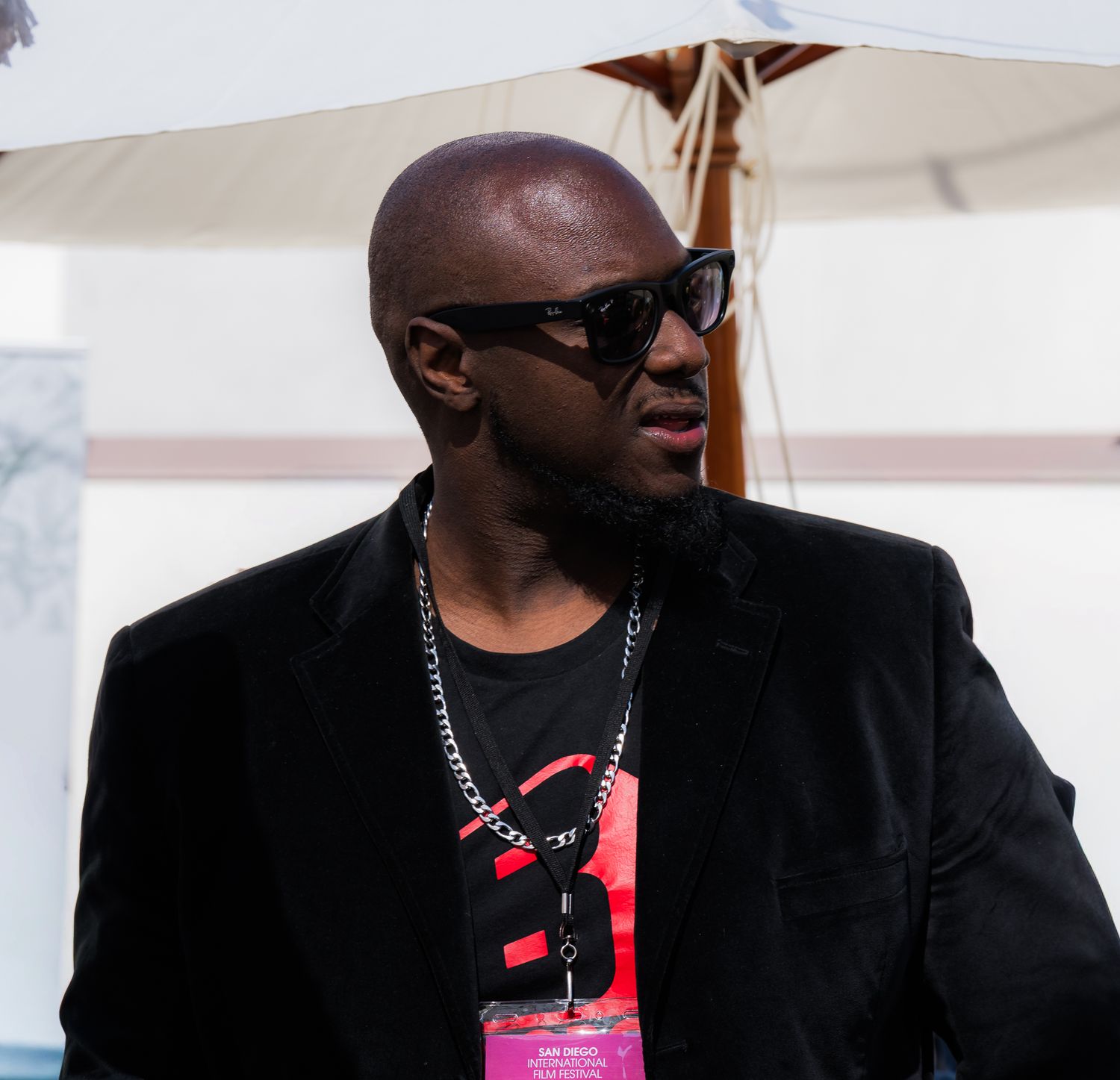 Bald man wearing black sunglasses, black blazer, and lanyard at an outdoor event.