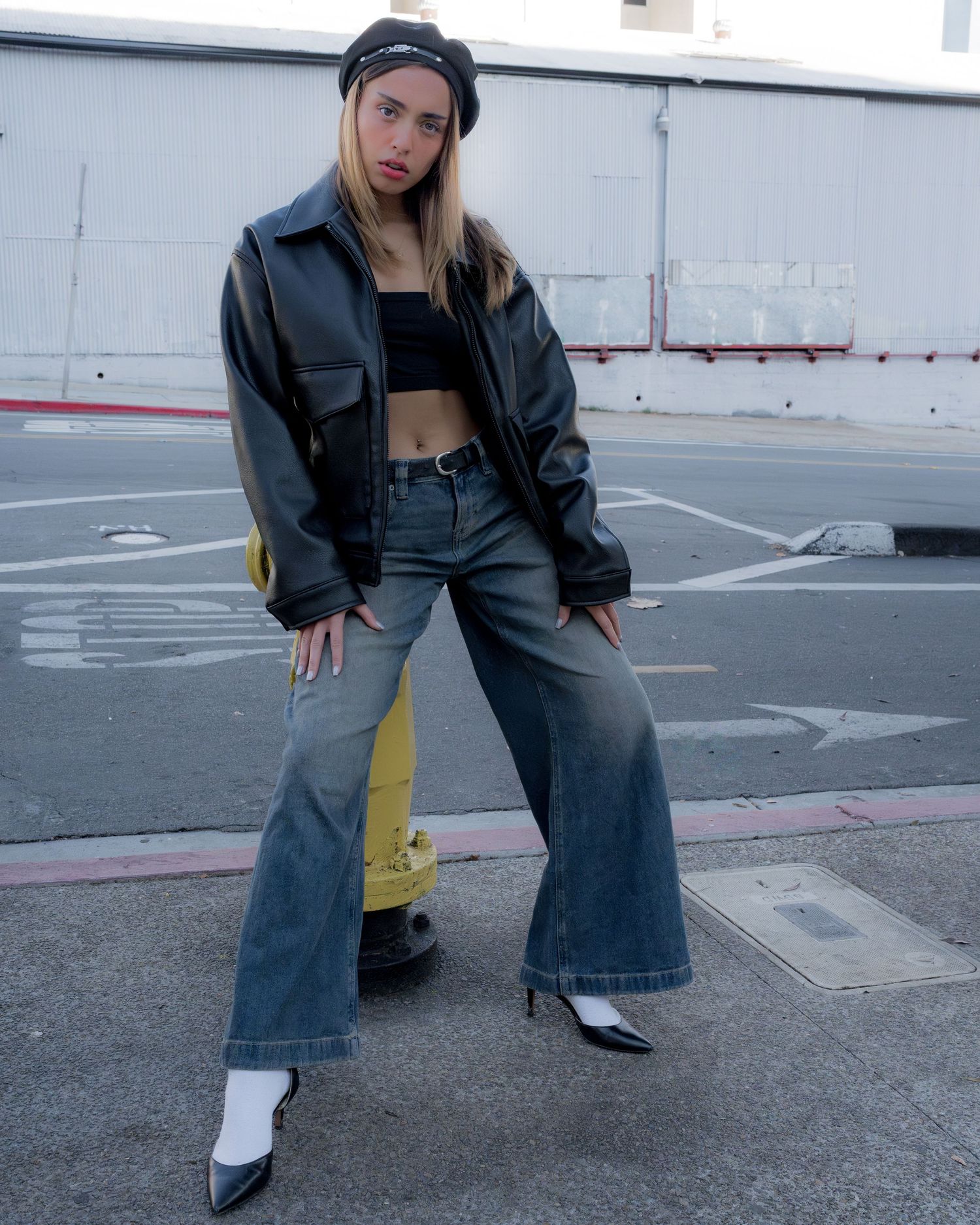Woman in black leather jacket, crop top, wide-leg jeans, and white boots poses on urban street.