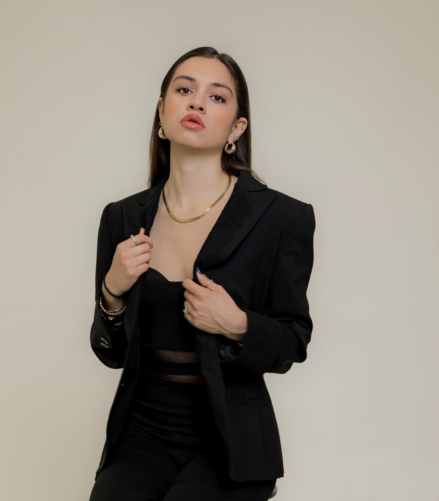 Young woman in black blazer and crop top with gold jewelry posing confidently against a neutral background.