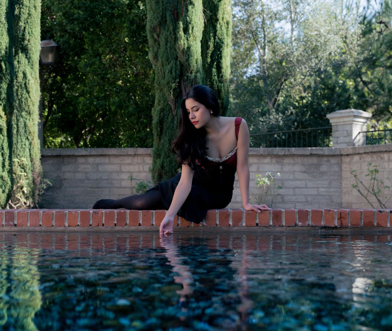 A woman in a black dress sits poolside, gently touching the water, surrounded by lush greenery and a brick wall.