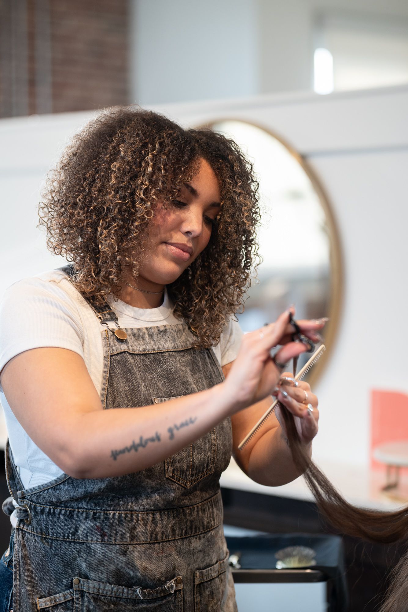Curly-haired hairstylist with tattoos cutting long hair in a modern salon with round mirrors.