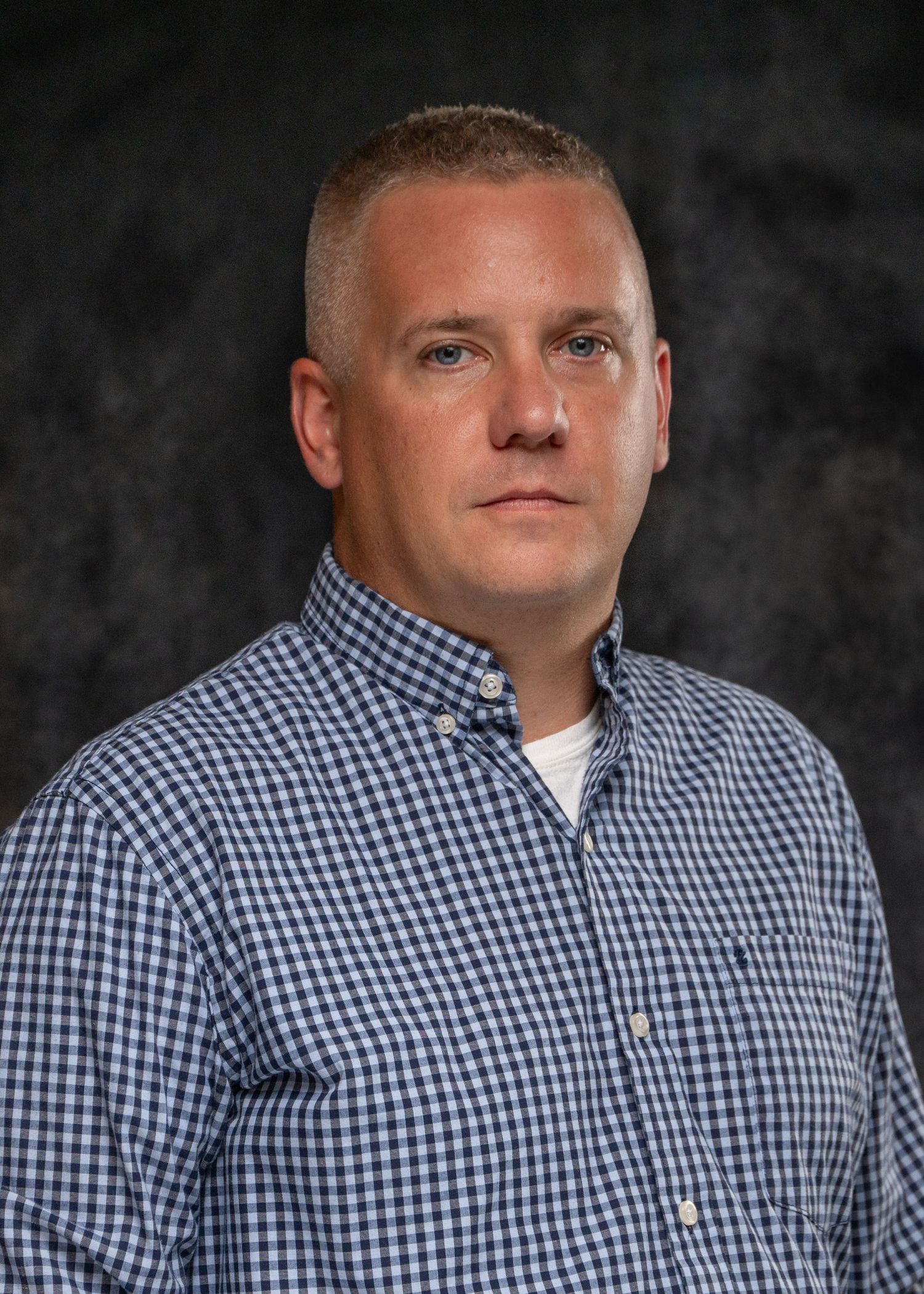Professional headshot of a man in a blue checkered shirt against a dark gray studio background.
