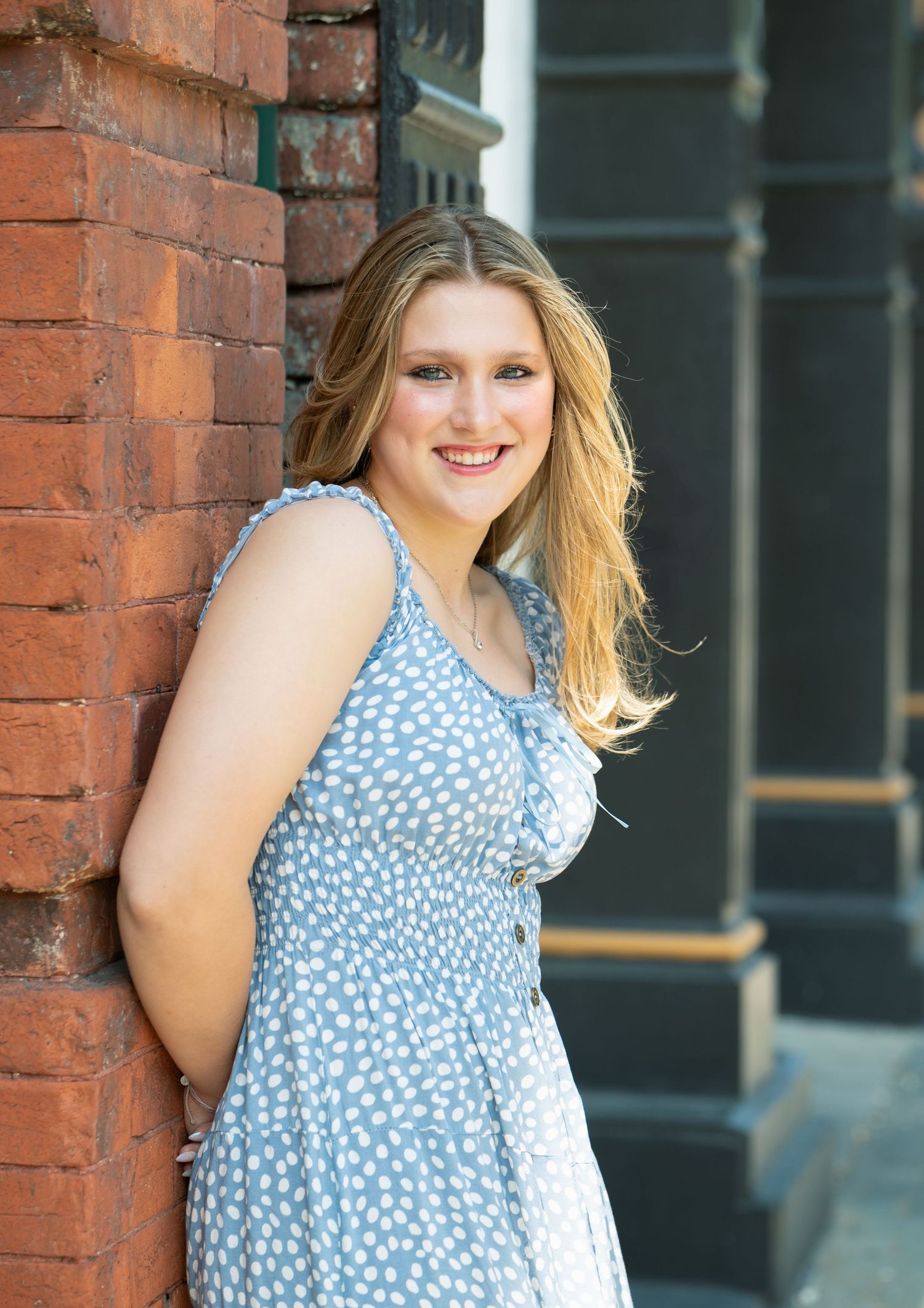 Young woman in blue floral sundress smiling against brick wall outdoors.