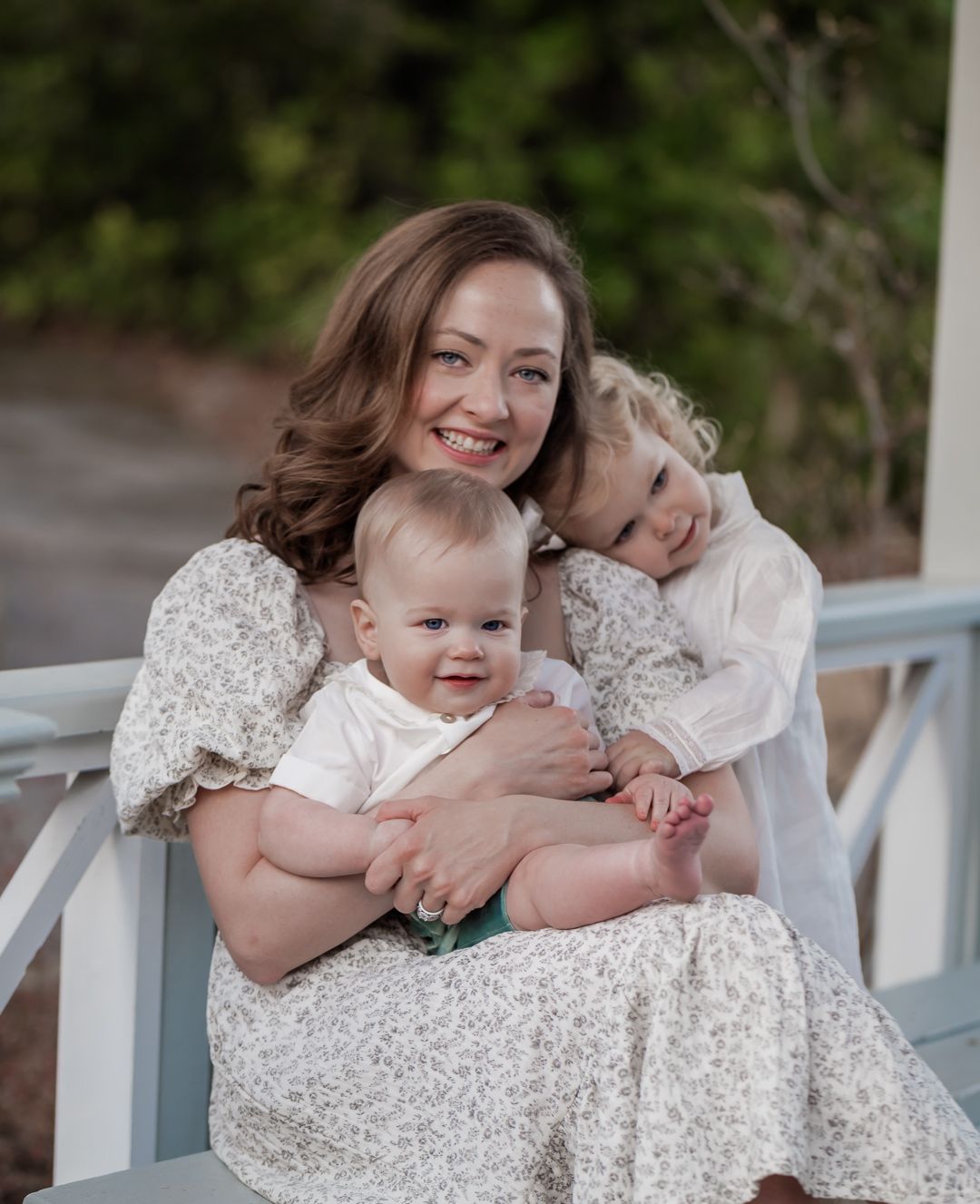 Smiling mother with wavy brown hair holding two young children on white porch in outdoor portrait.