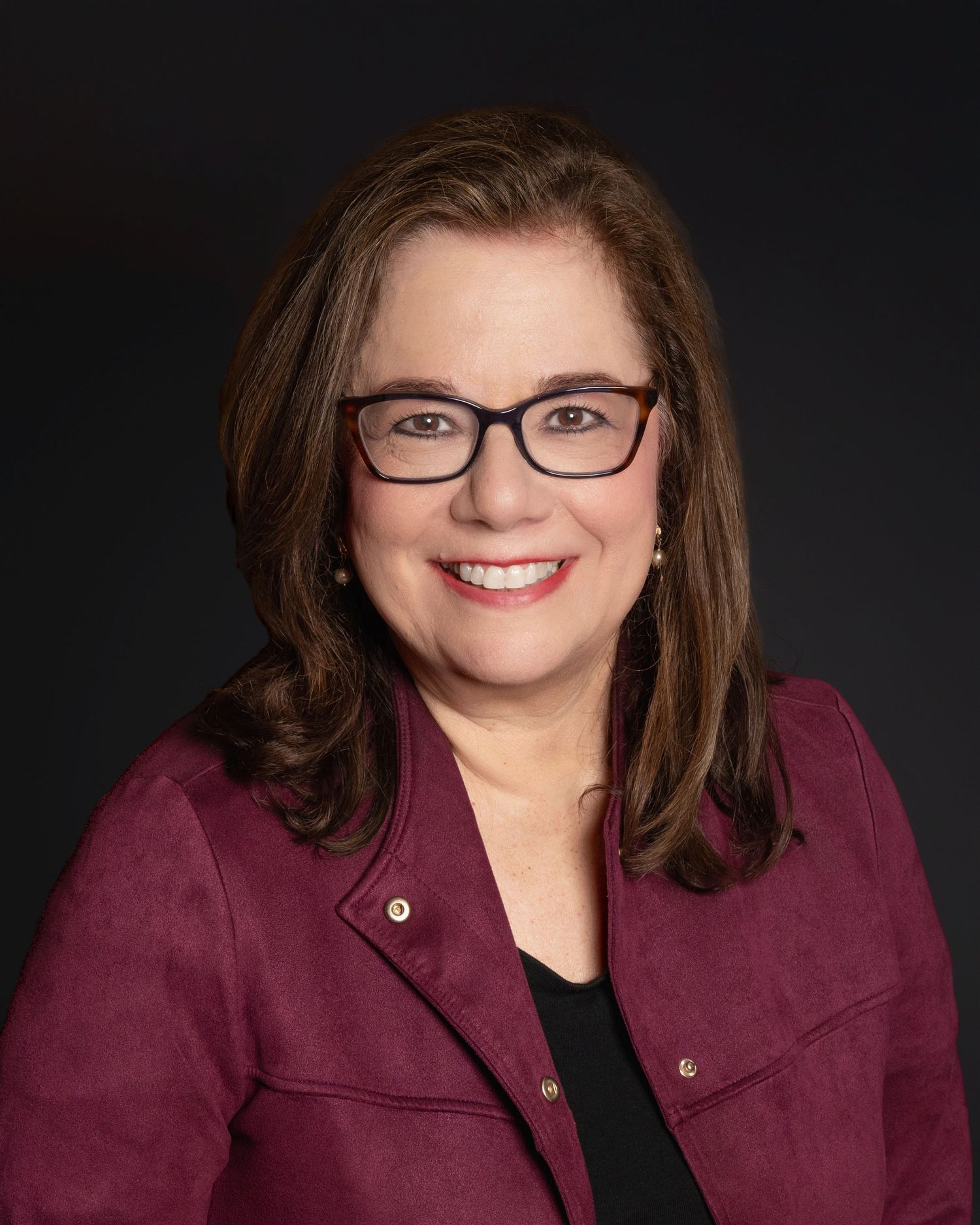 Smiling woman with brown hair and glasses wearing a burgundy jacket against dark background.