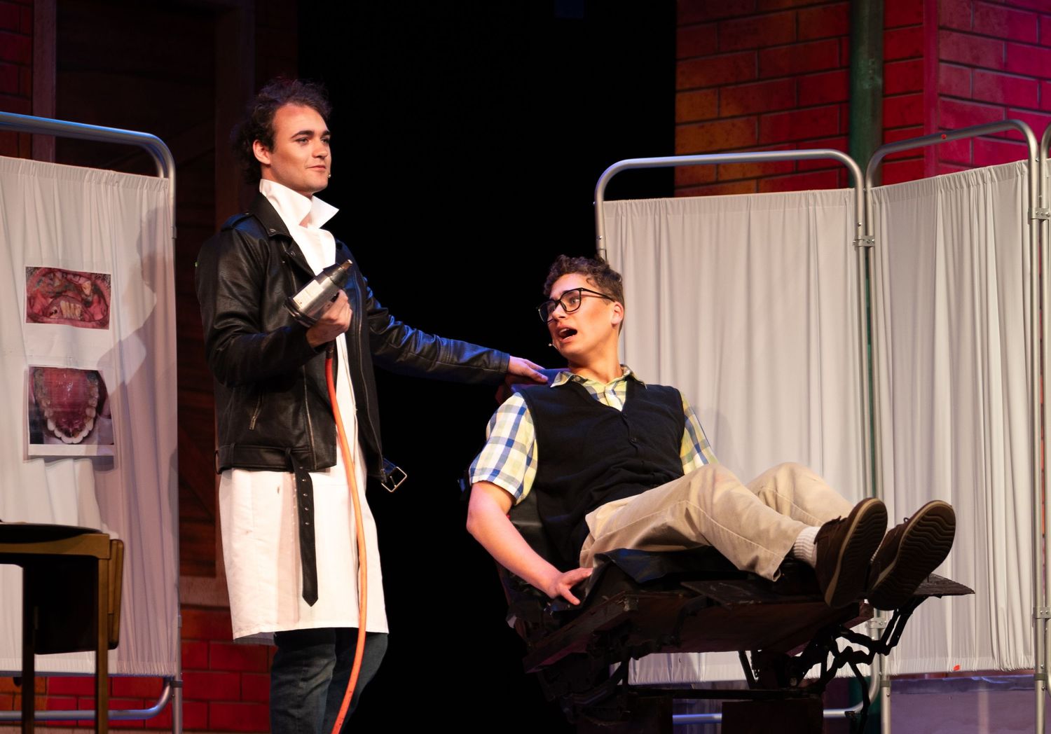 Two actors perform a dental scene on stage, one in a leather jacket holding a tool over a patient in a chair.