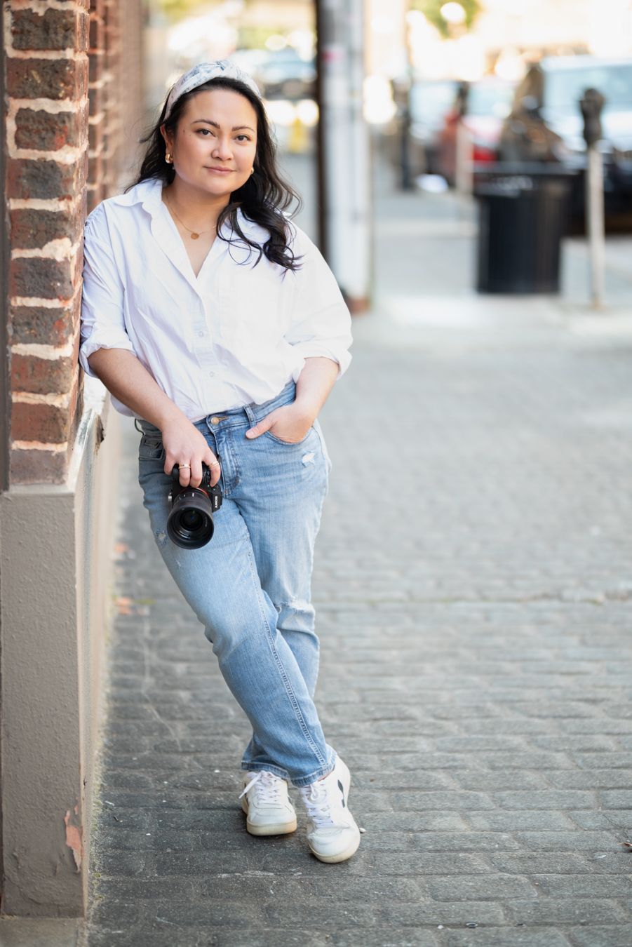 Female photographer posing confidently on a city sidewalk holding a professional camera.