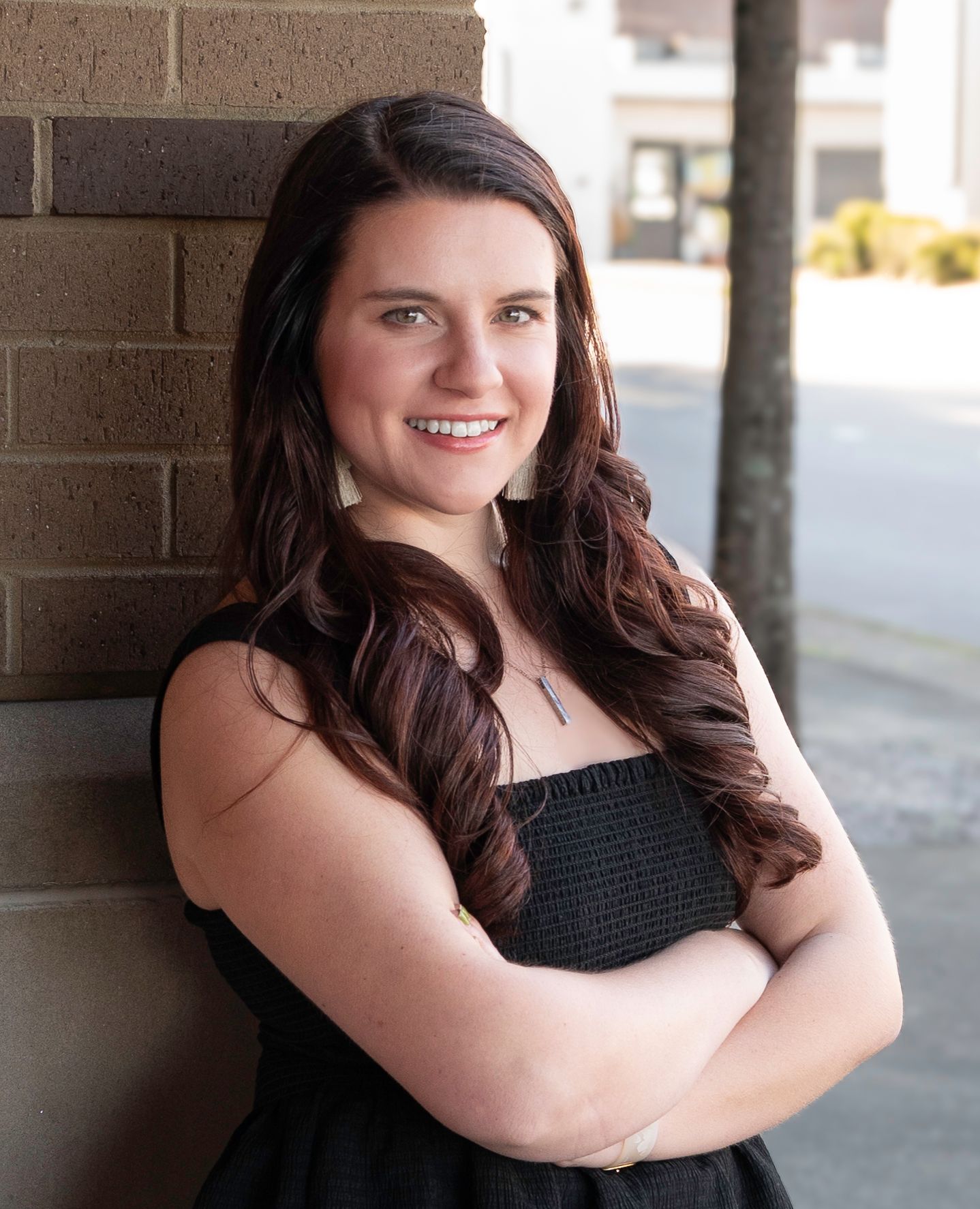 Young woman with long brown hair smiling, arms crossed, leaning against brick wall outdoors in black dress.