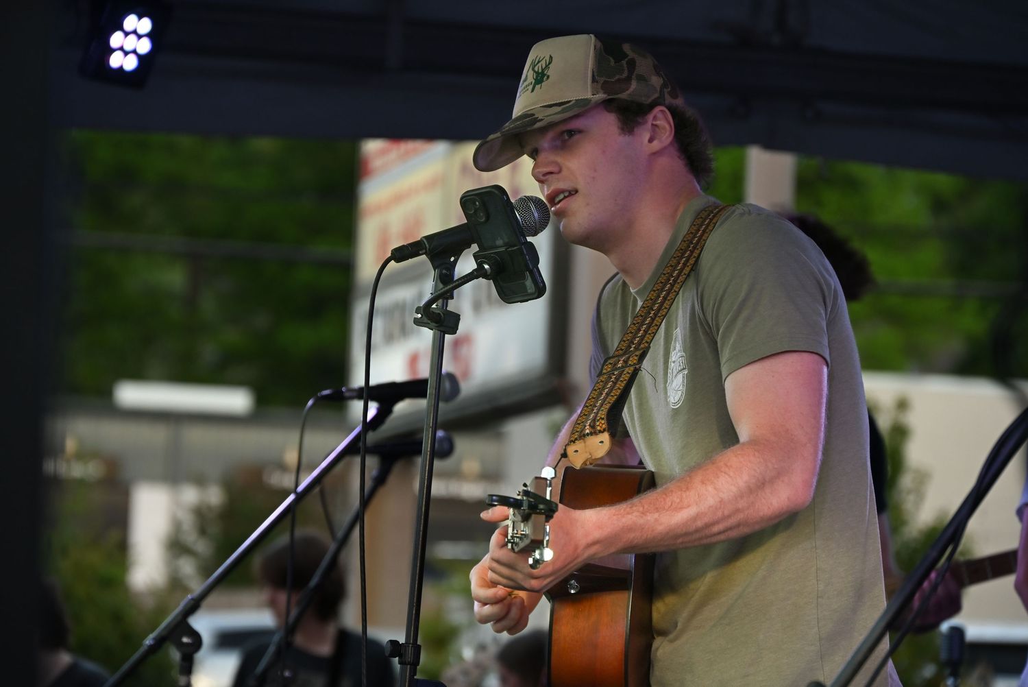Male musician in camo cap playing acoustic guitar and singing into microphone at outdoor daytime concert.