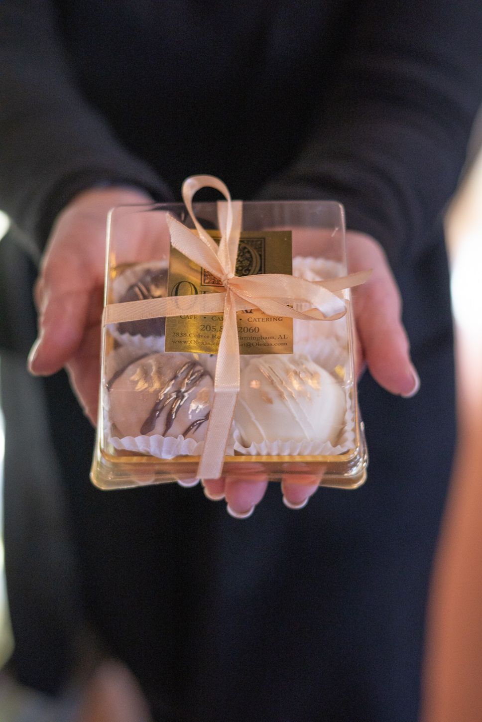 Hands holding a clear gift box with macarons tied with a pink ribbon bow and gold label.