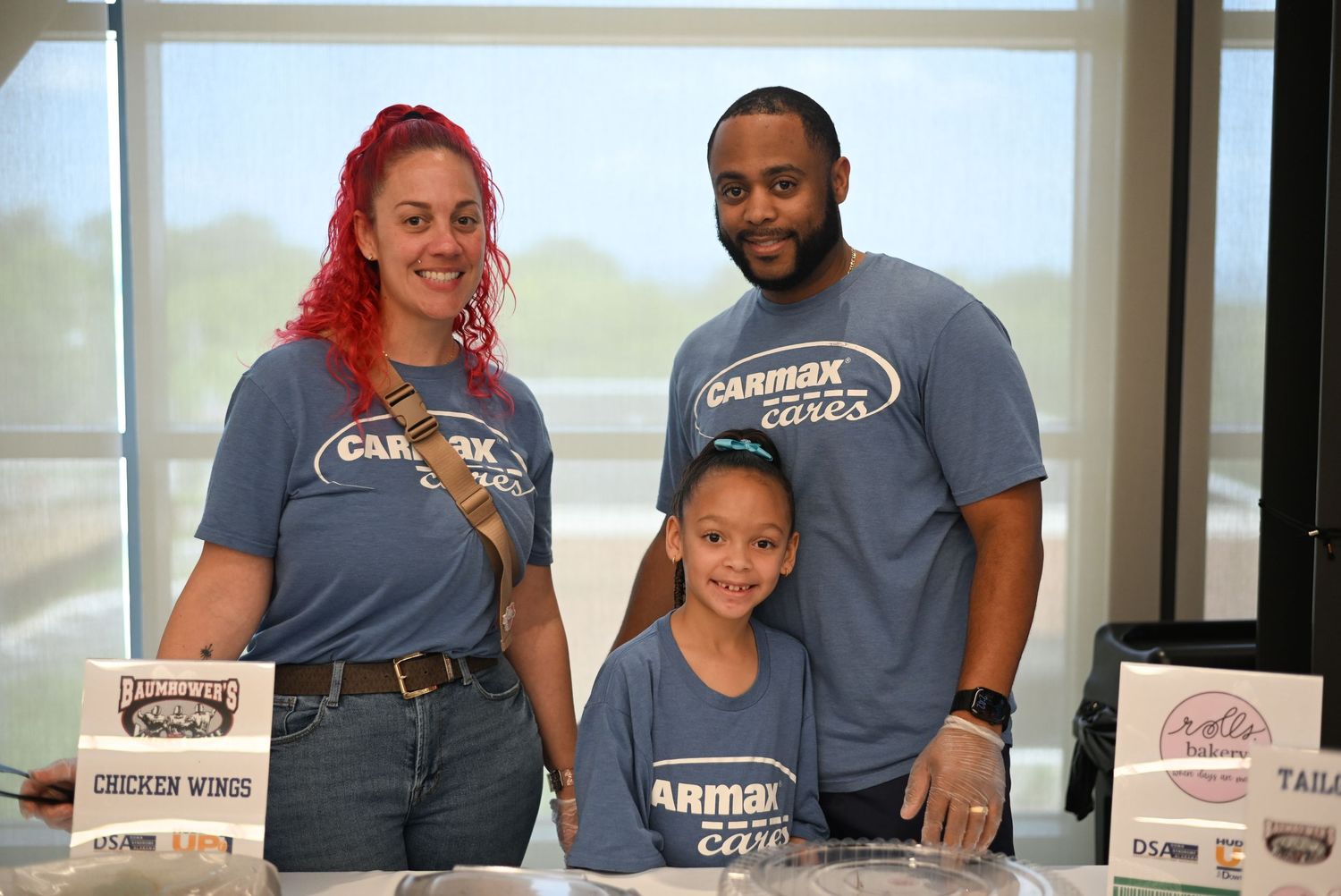 Family of three wearing CarMax Cares t-shirts smiling at a community event booth with food signs.