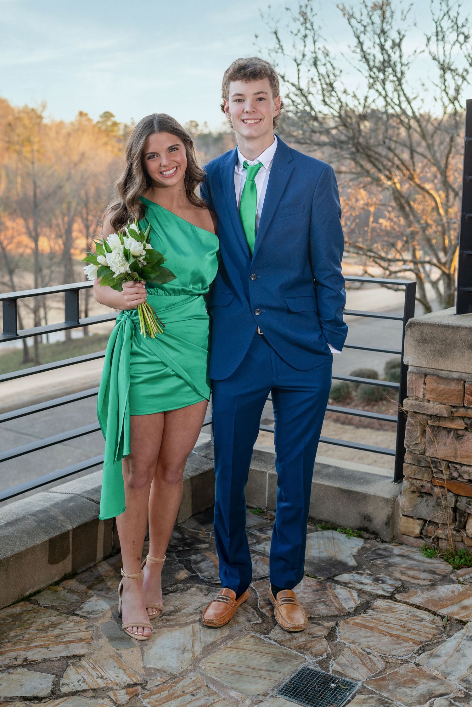 Young couple dressed for a formal, girl in green dress holding white flowers, boy in blue suit with green tie, outdoors.