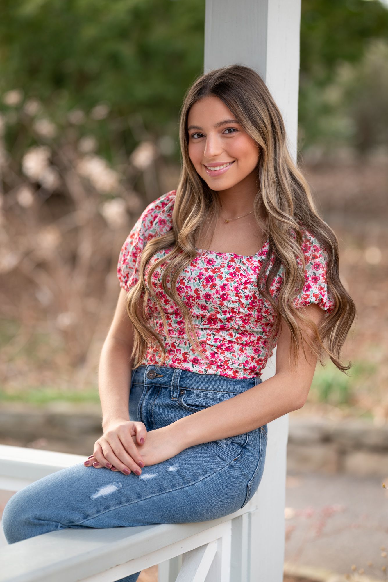 Young woman with long blonde hair smiling in a floral top and jeans outdoors.