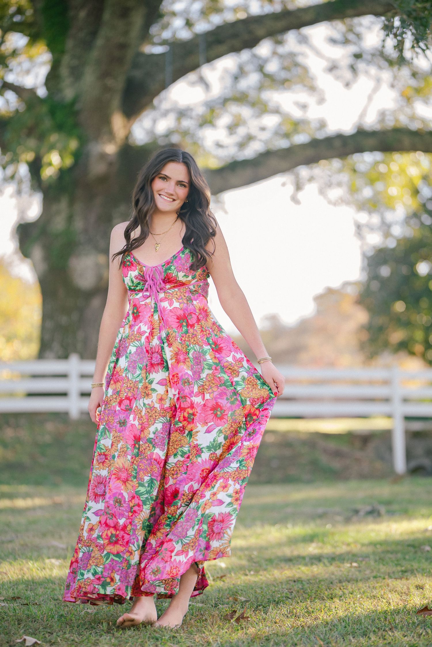Brunette woman in pink floral maxi dress twirling under large oak tree by white fence outdoors.