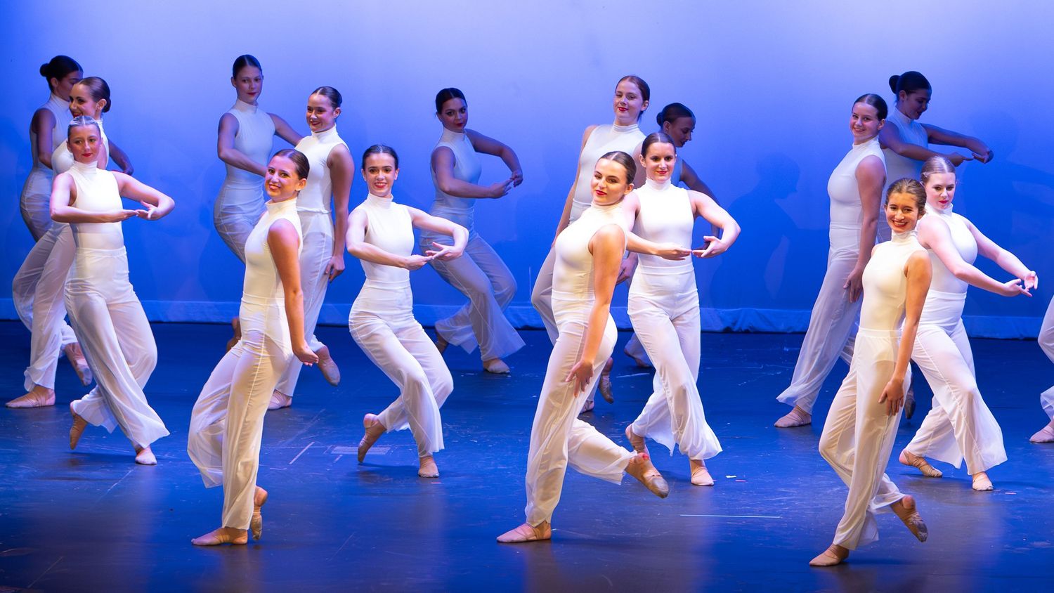 Dancers in white costumes perform a synchronized routine on a blue-lit stage during a dance recital.