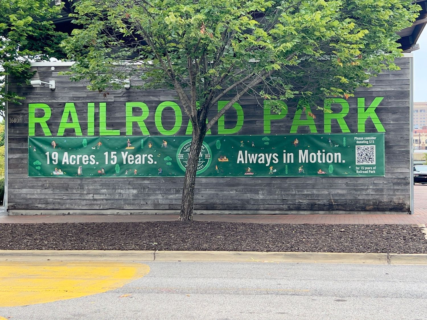 Railroad Park banner sign reading '19 Acres. 15 Years. Always in Motion.' displayed on a brick wall in Birmingham, AL.