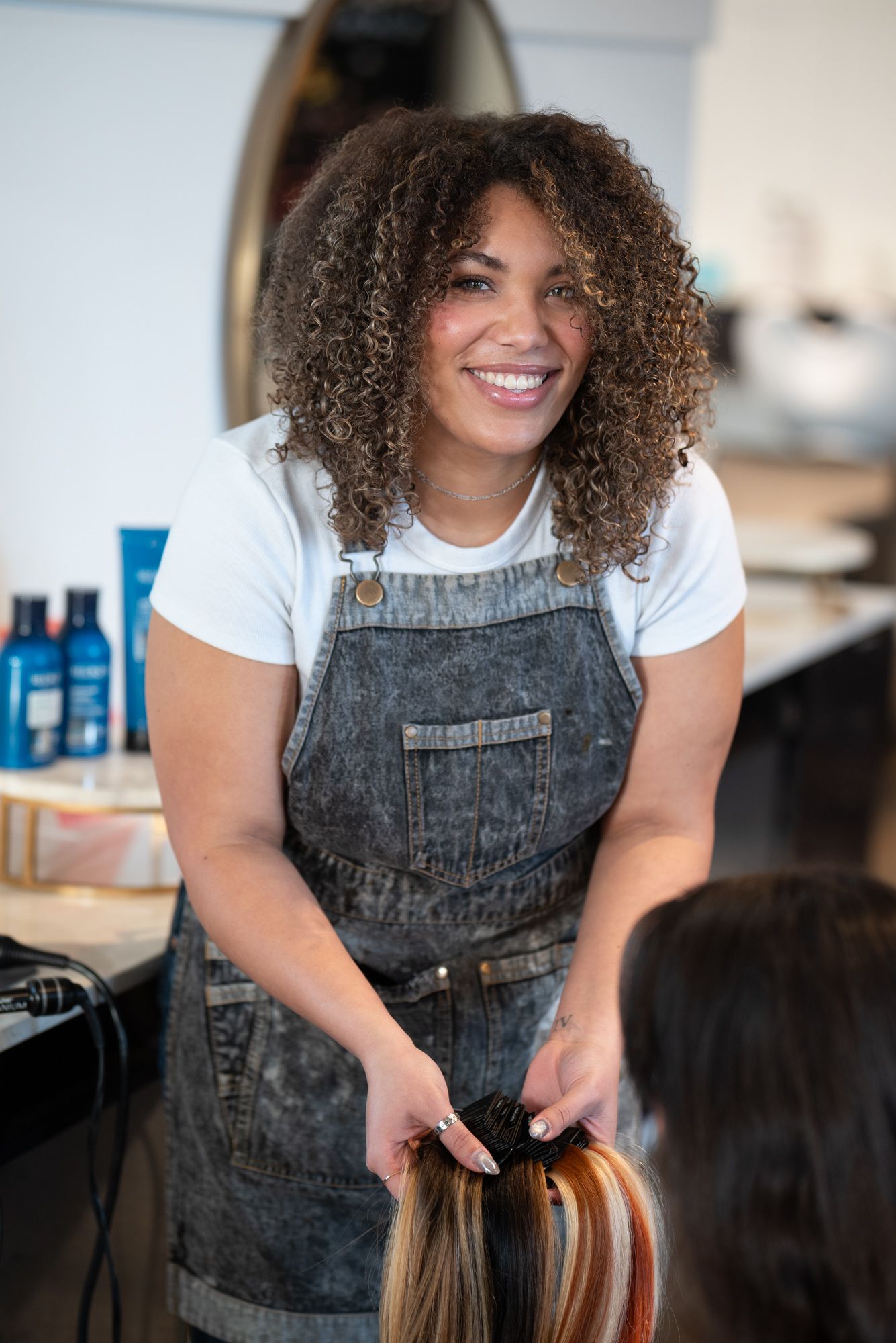 Professional hairstylist in denim overalls works on client's hair in modern salon setting in Birmingham, AL.