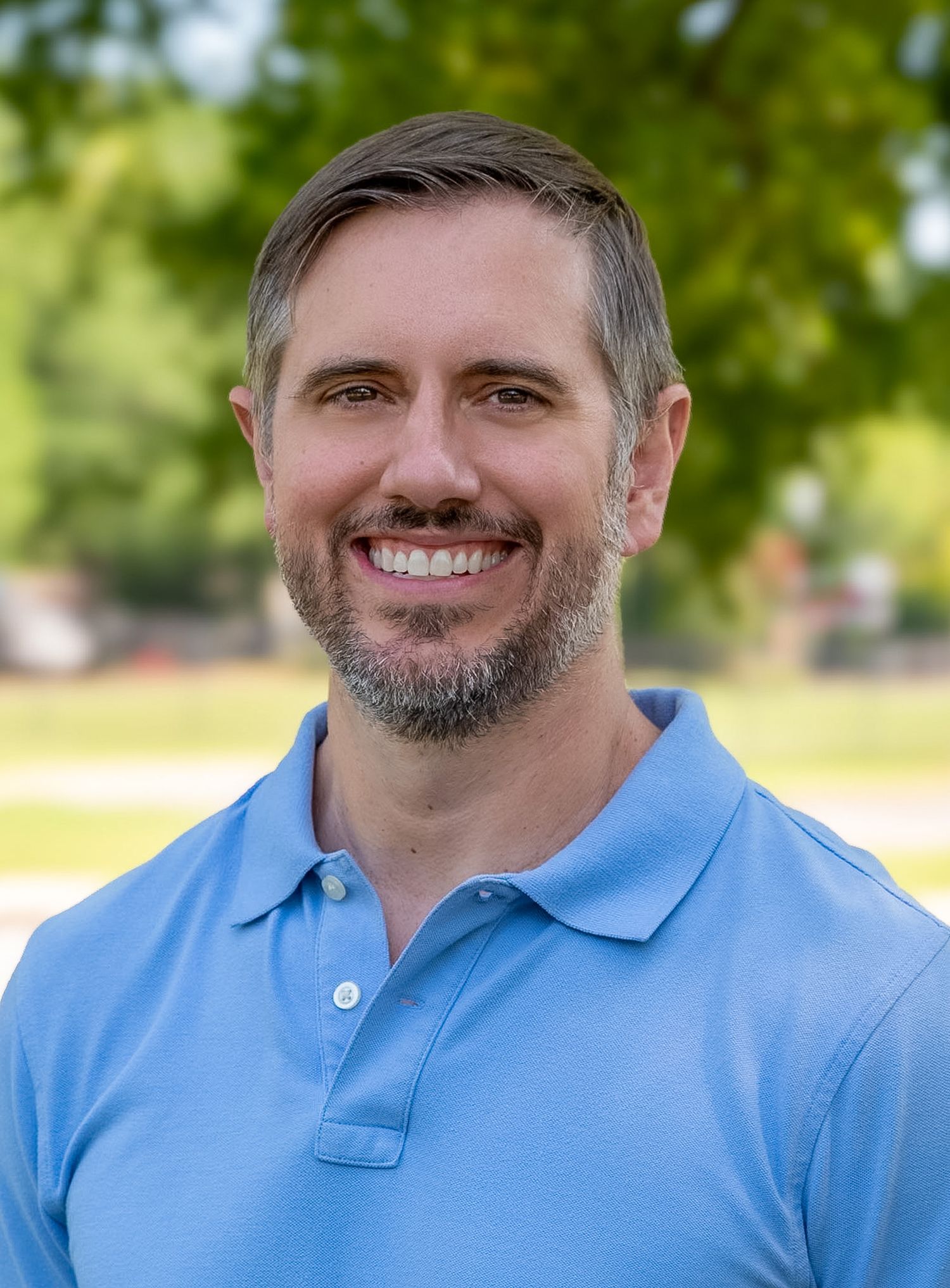 Smiling man with short hair and beard wearing a light blue polo shirt outdoors.