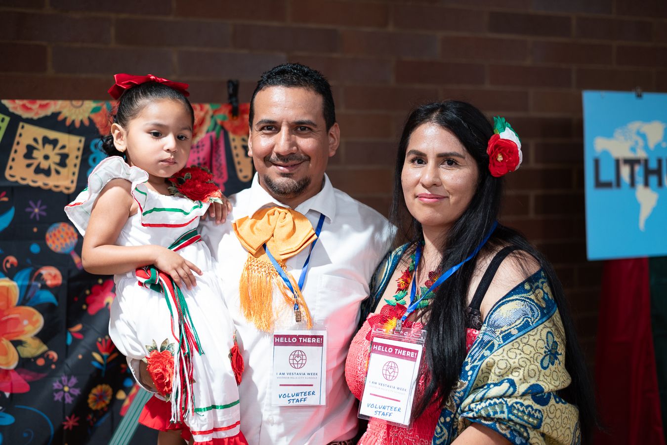 Hispanic family of three posing at a cultural event, man wearing orange medal, woman with red flower in hair.