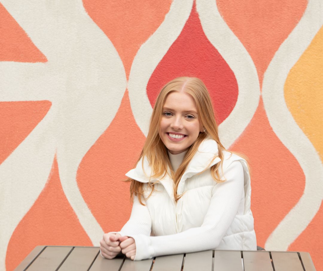 Smiling young woman with long red hair wearing a white vest, seated at a wooden table against a colorful orange wave mural.