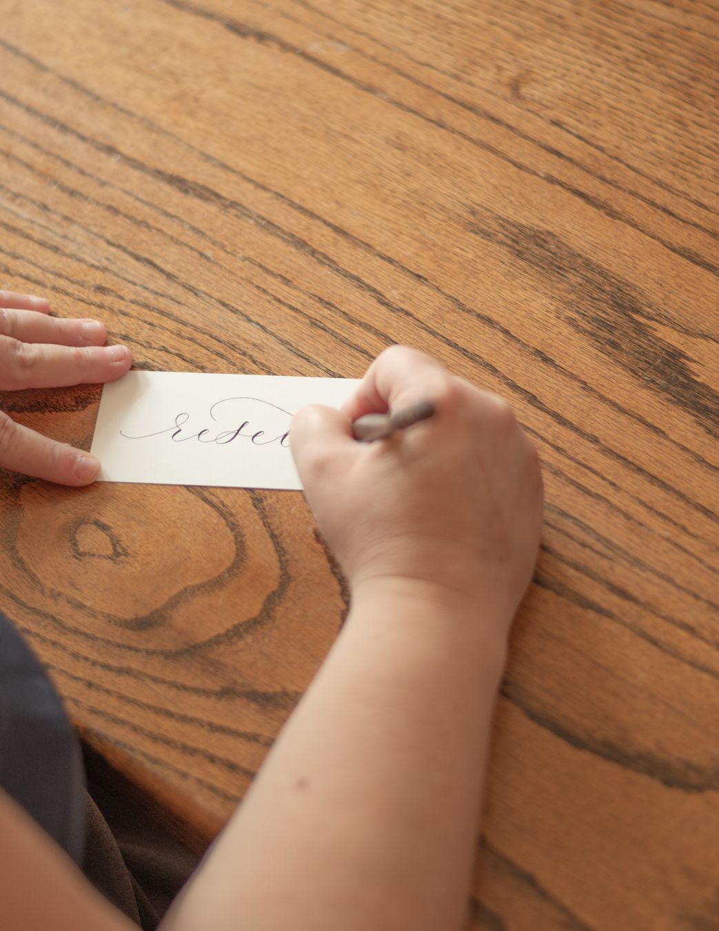 Person practicing calligraphy on a small white card at a wooden table, writing elegant cursive script with a pen.