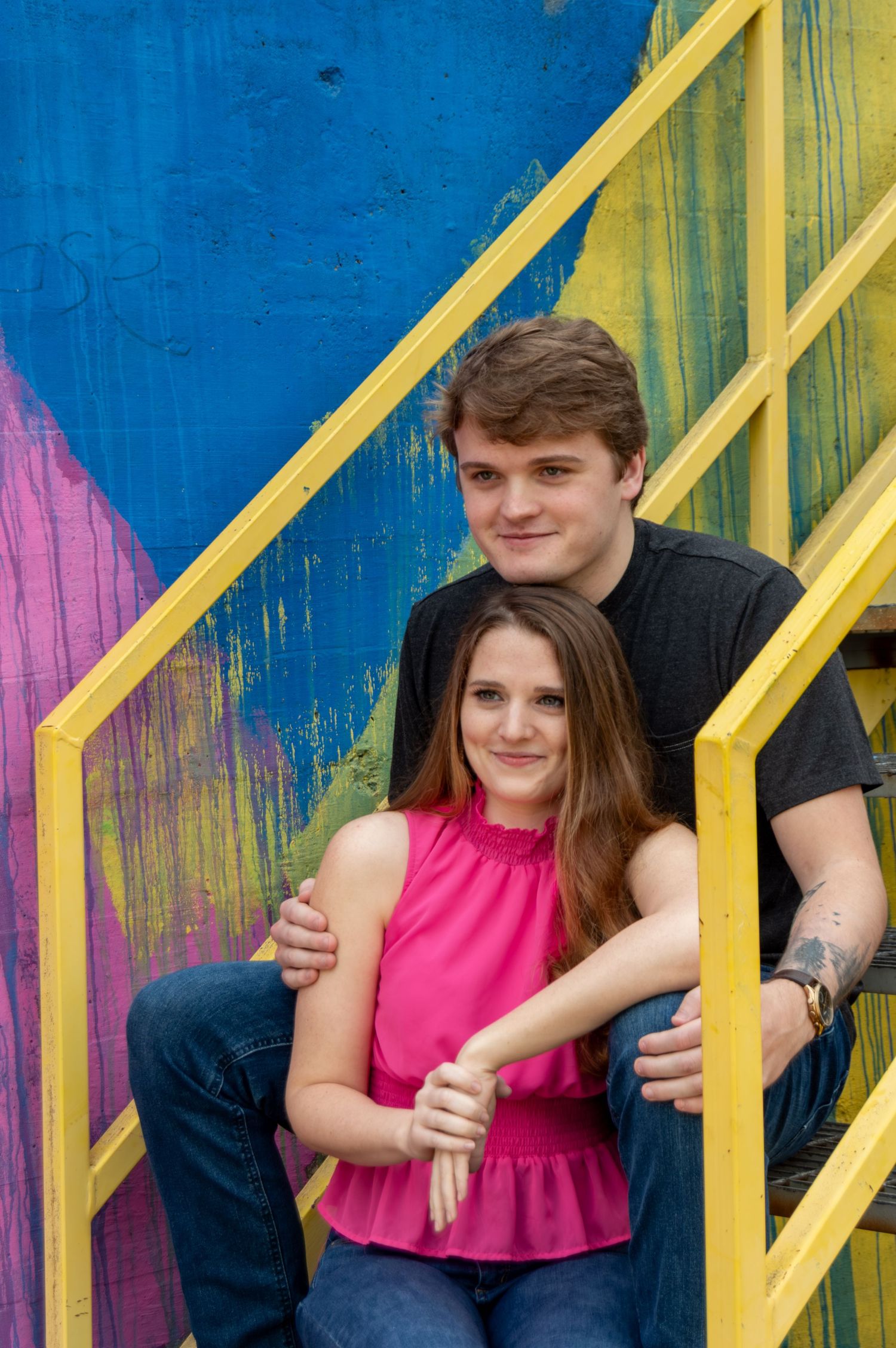 Young couple posing on colorful painted stairs, woman in pink top sitting in front of man in black t-shirt.