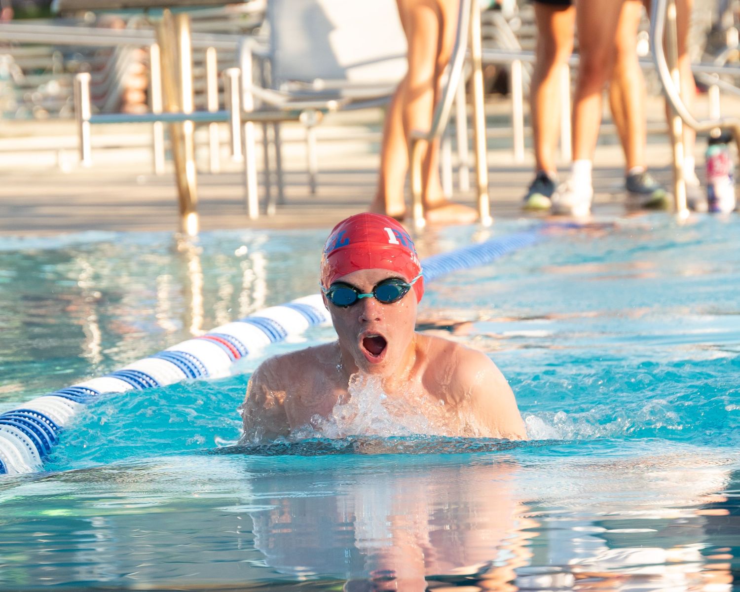 Young competitive swimmer in red cap and goggles performs breaststroke in an outdoor pool during a swim meet.