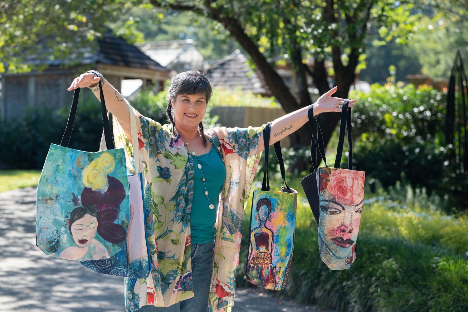 Woman displaying colorful artistic tote bags with painted faces outdoors in a garden setting.