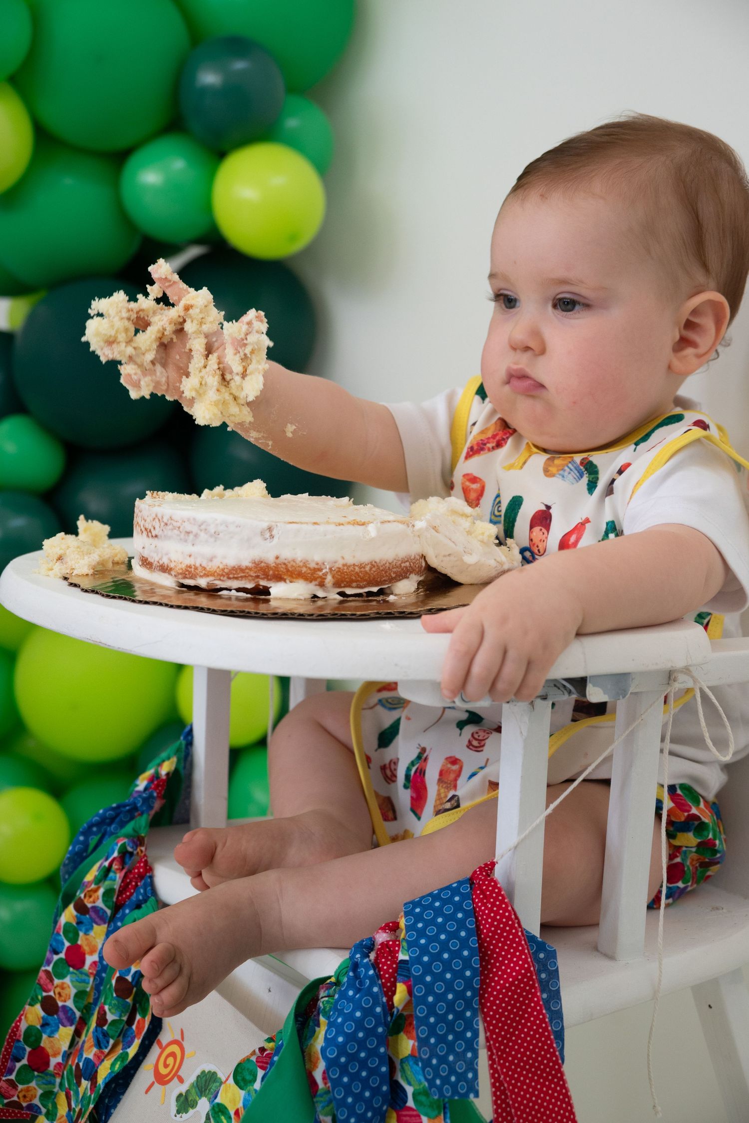 Baby in high chair smashing first birthday cake, surrounded by green and yellow balloons at a first birthday celebration.
