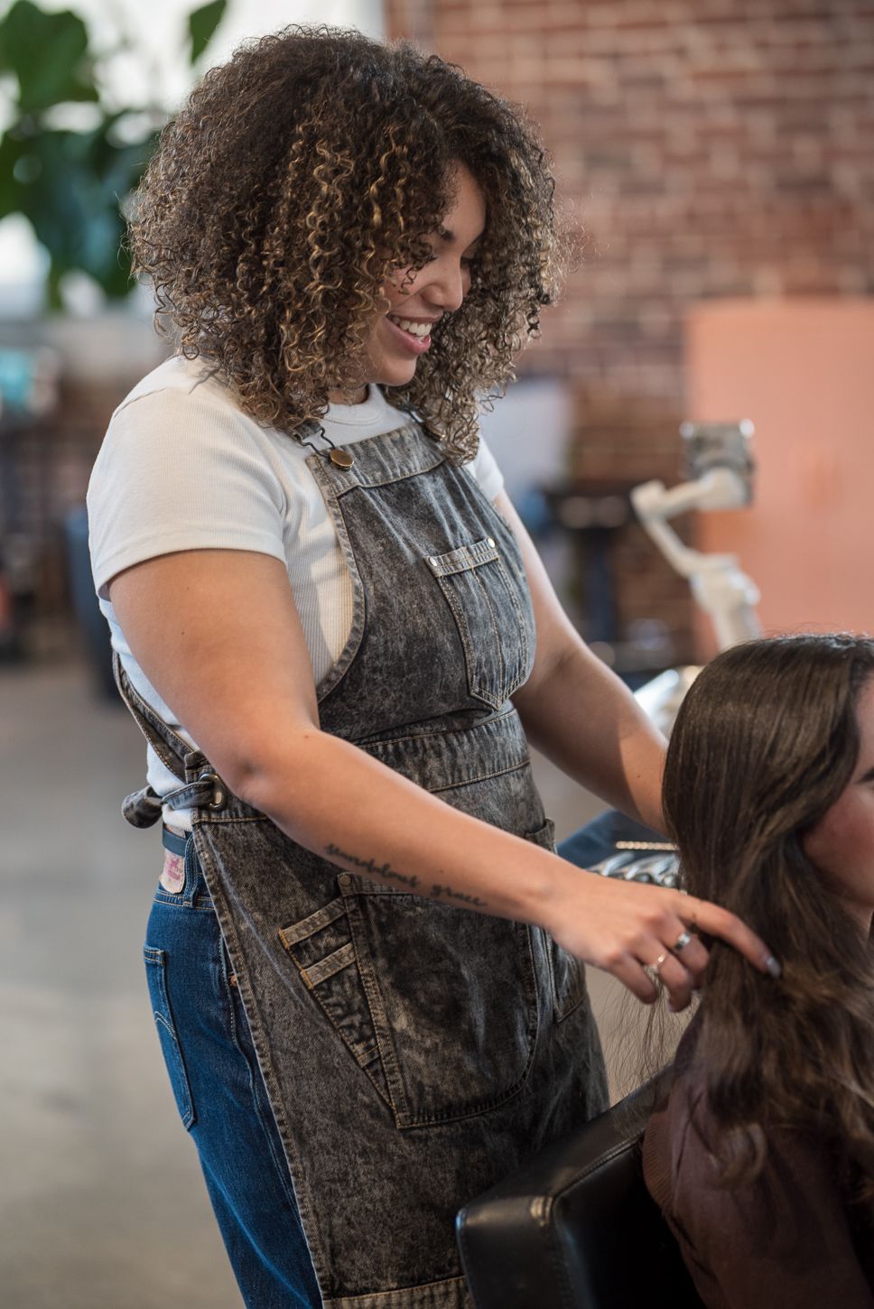 Curly-haired hairstylist in denim apron smiling while styling client's hair in modern salon.