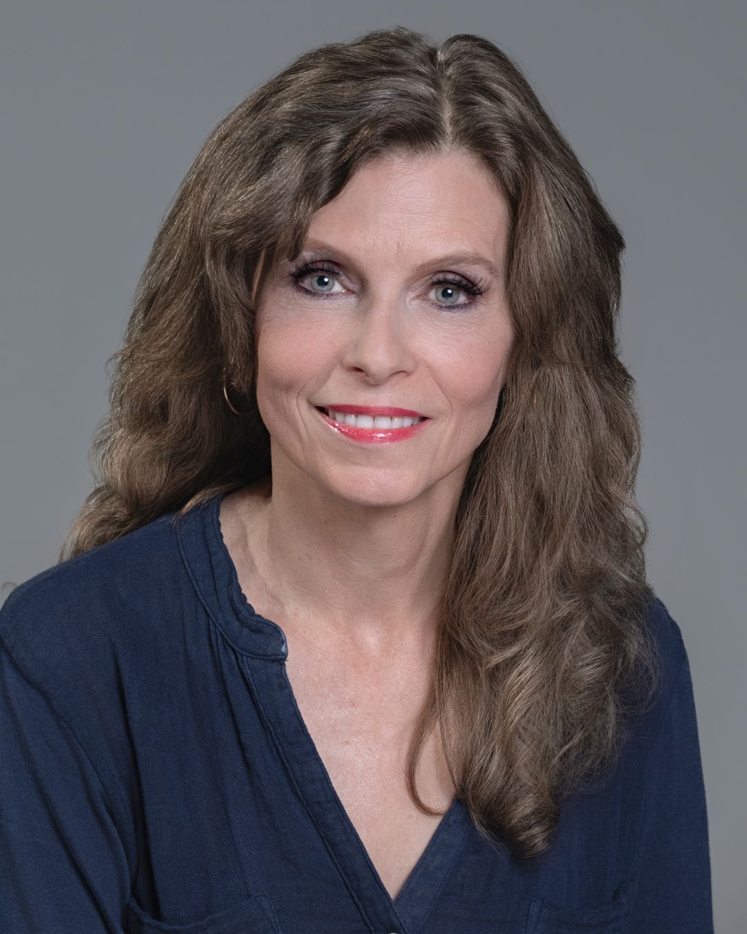 Professional headshot of a middle-aged brunette woman in a navy blouse on gray background.