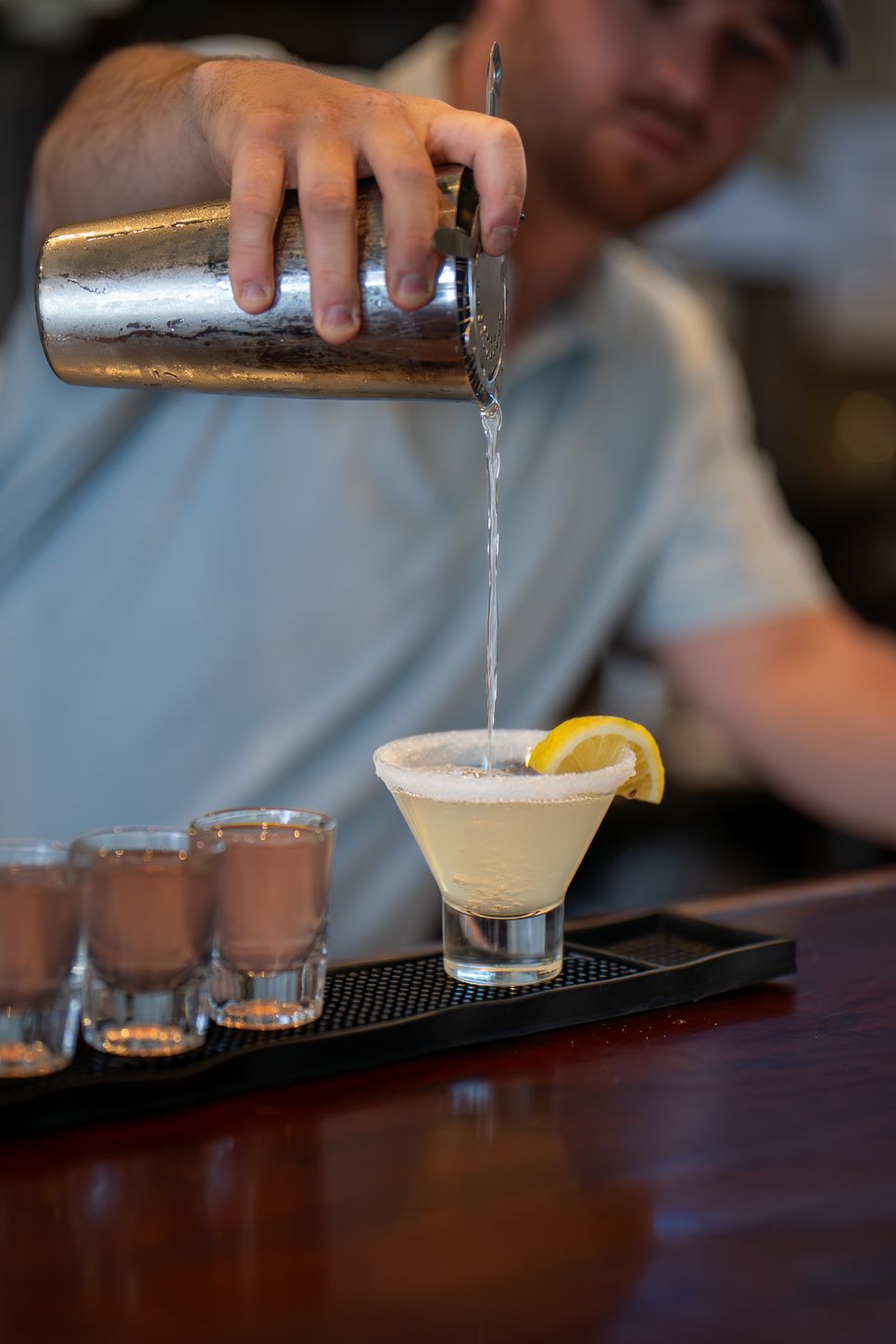 Bartender pouring a frothy cocktail into a martini glass garnished with lemon on a bar tray with shot glasses.