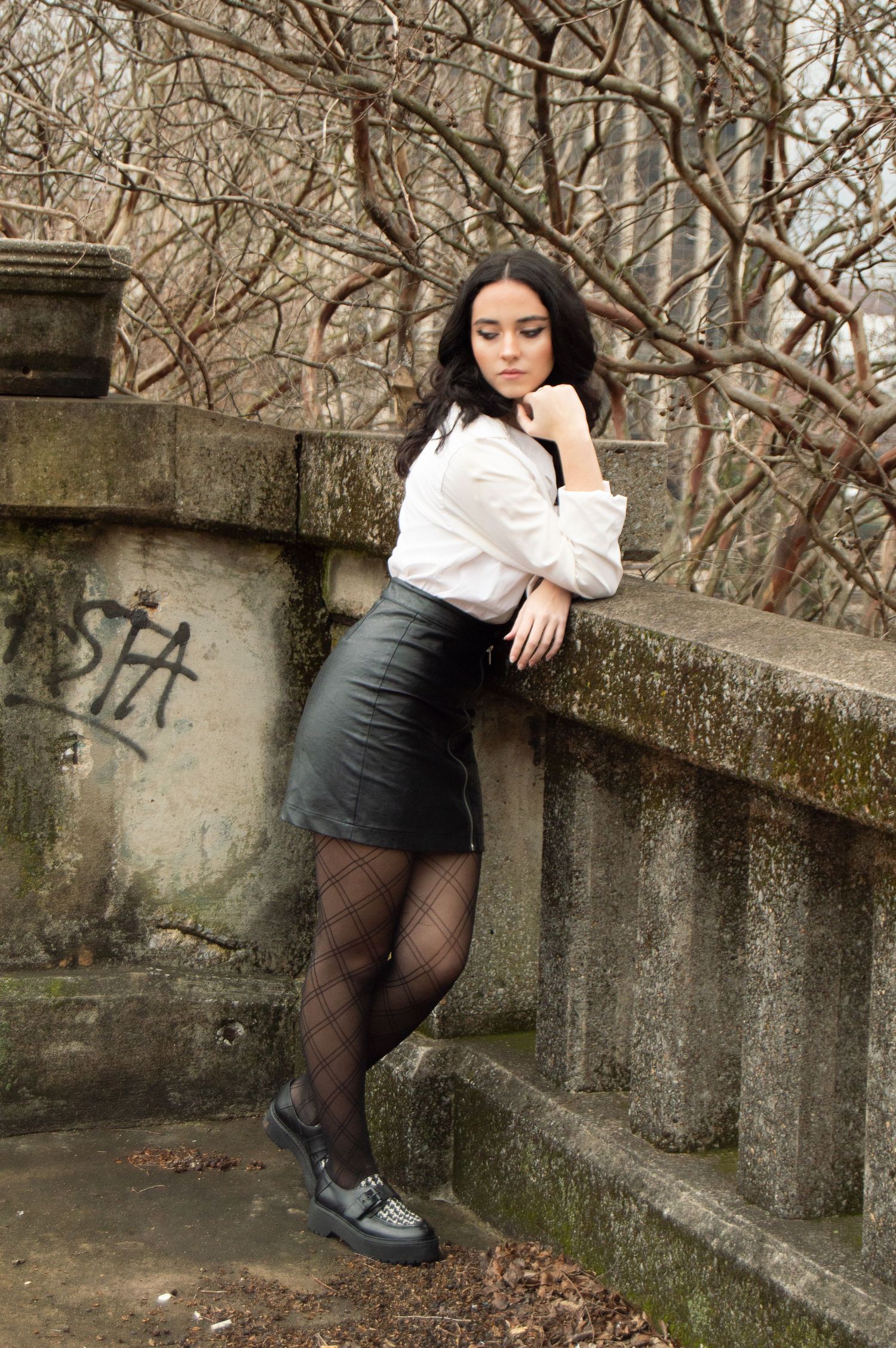 Young woman in white blouse and black leather mini skirt posing against stone wall in urban outdoor setting.