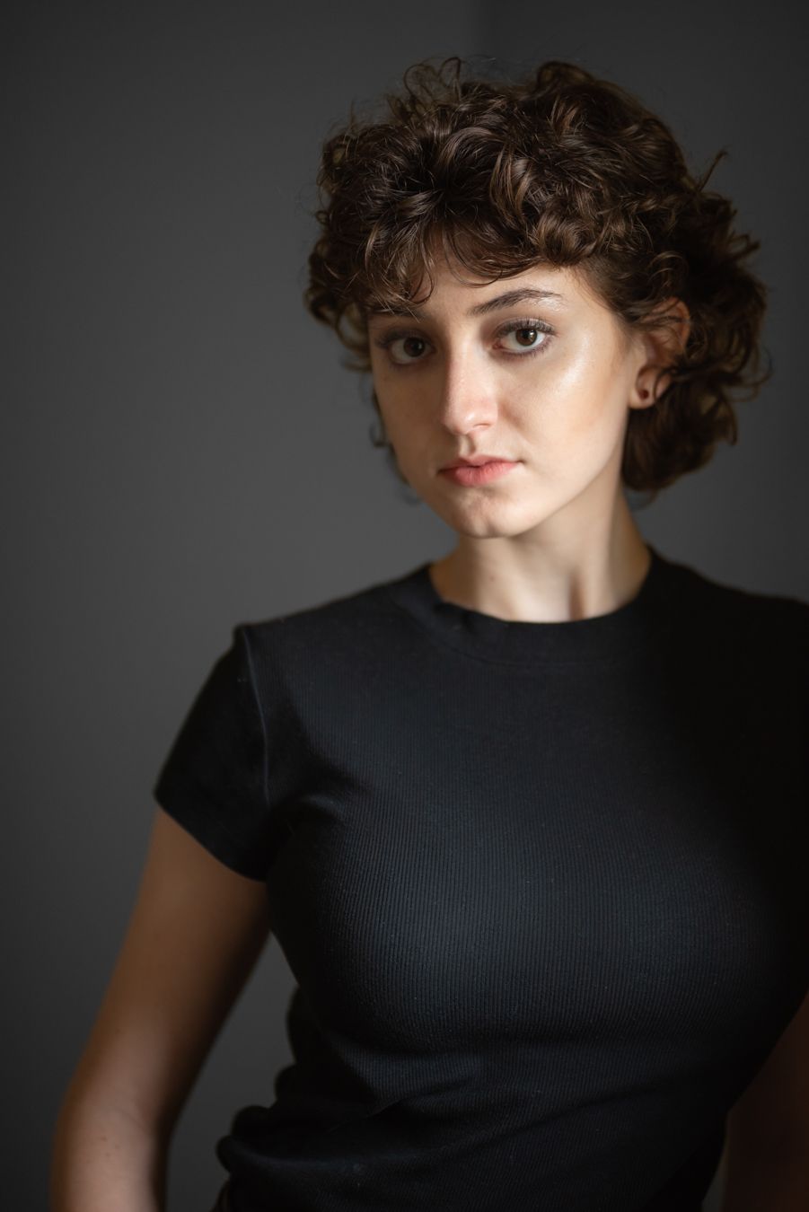Young woman with short curly hair wearing a black t-shirt poses against a dark gray background.