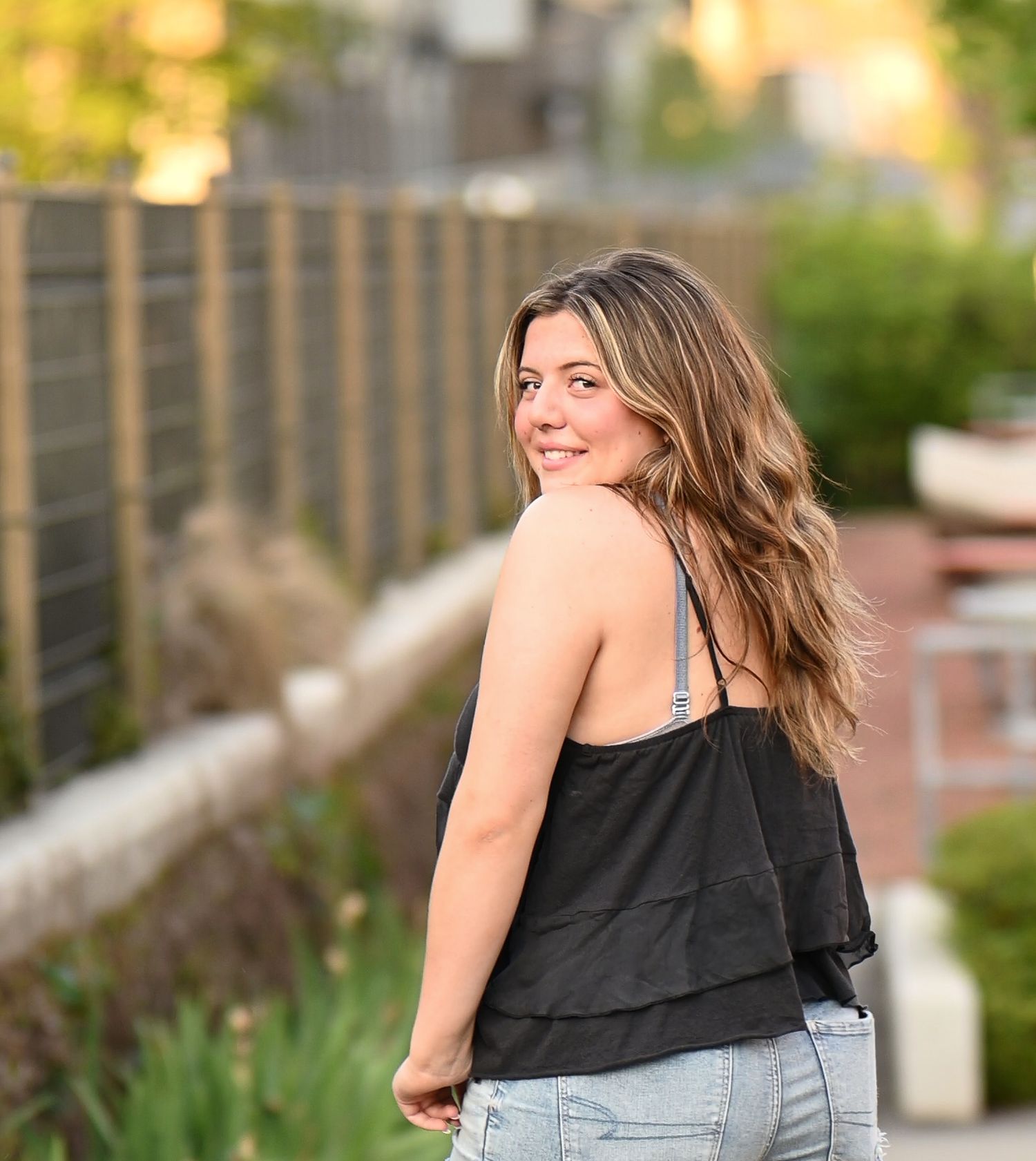 Young woman in black tank top and jeans smiling over shoulder outdoors.
