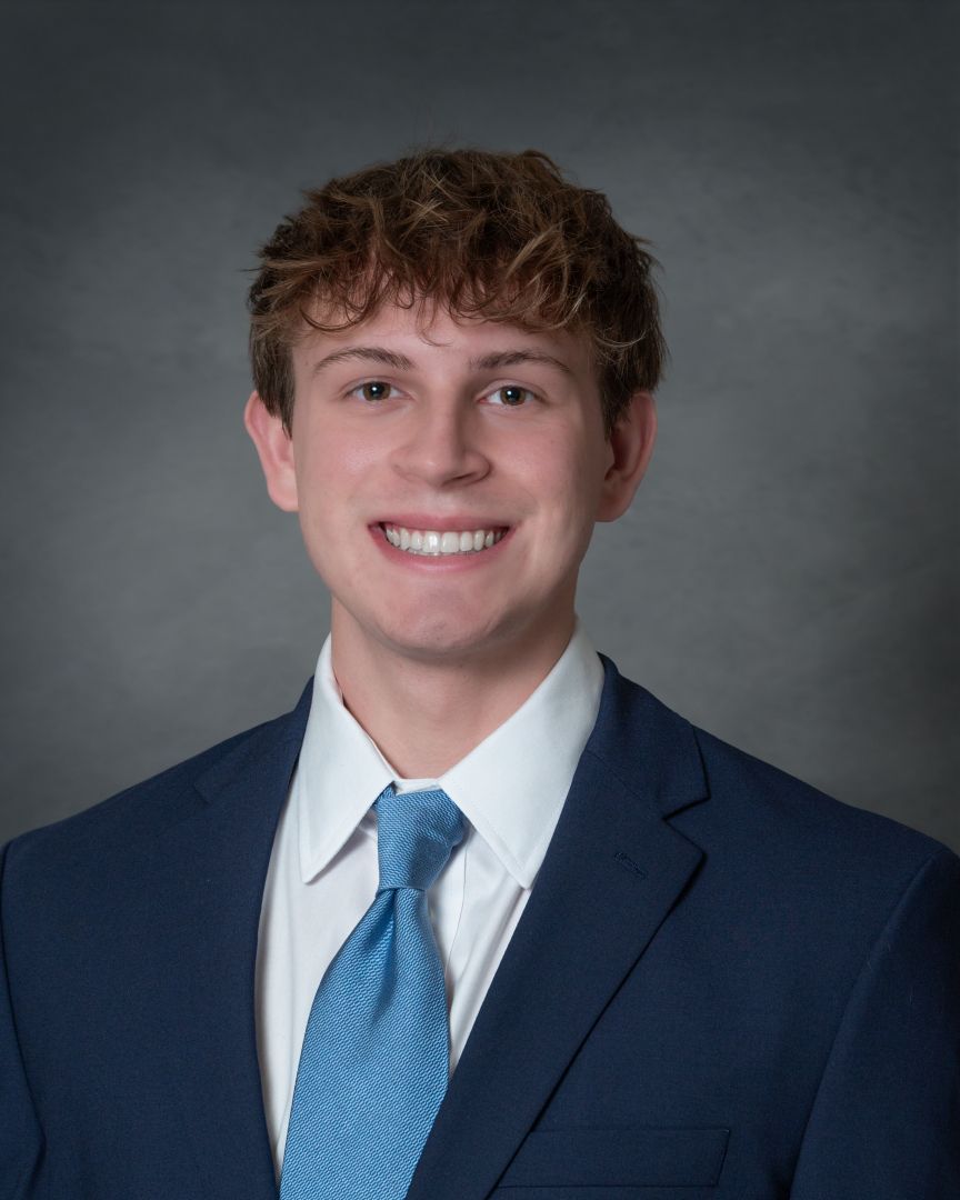 Young man in navy blue suit and light blue tie smiling for a professional headshot on gray background.