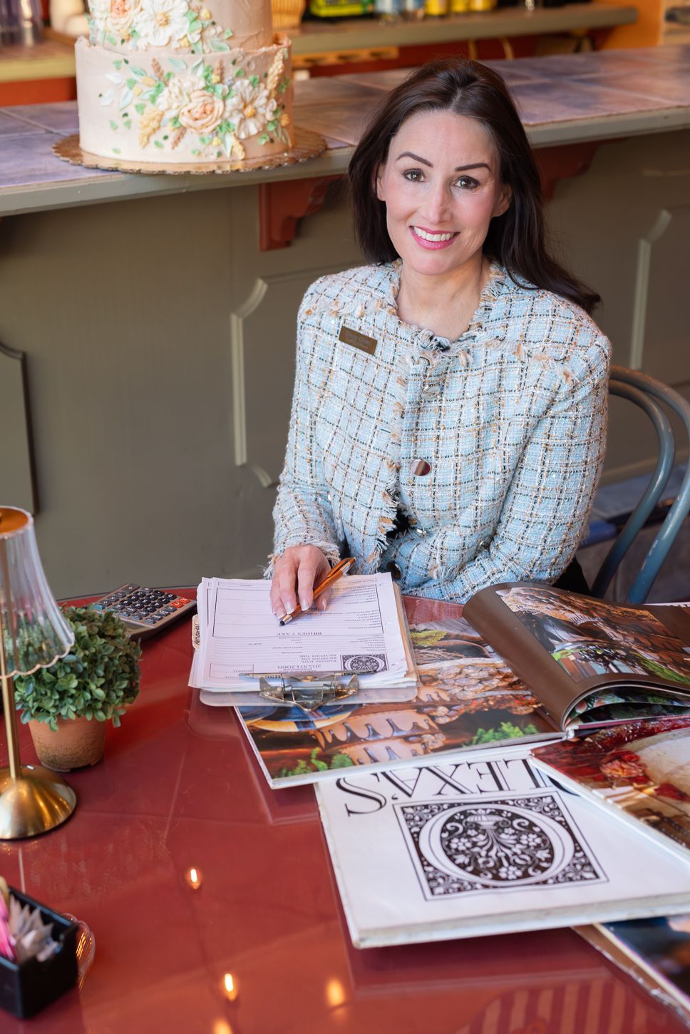 A smiling woman in a blue tweed jacket sits at a red table with open books and a decorated cake in the background.