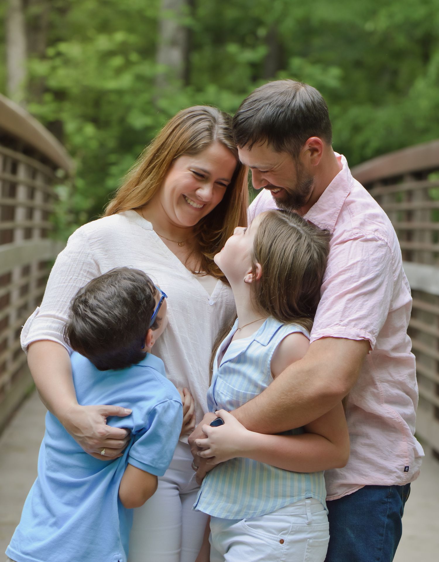 Happy family of four sharing a warm embrace on a wooden bridge surrounded by green trees.