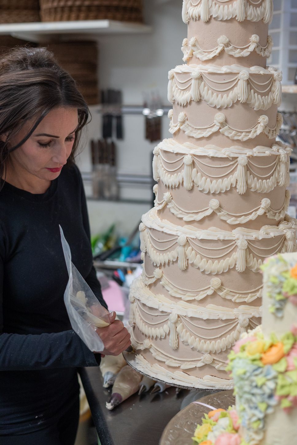 Woman in black decorating an elegant multi-tiered wedding cake with white frosting details.