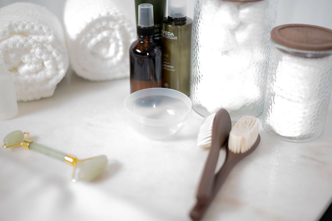 Skincare tools including jade roller, facial brush, oils and cotton pads on marble surface.
