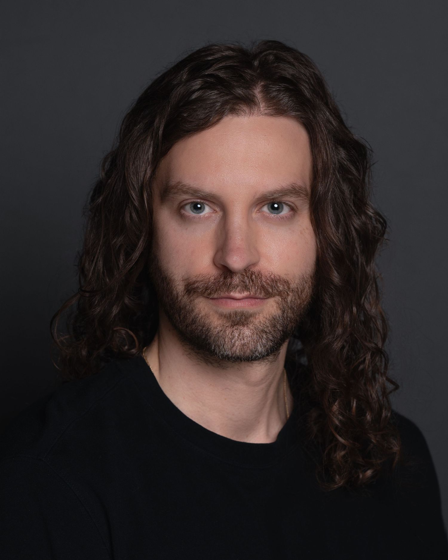 Professional headshot of man with long curly brown hair and beard wearing black shirt against gray background.
