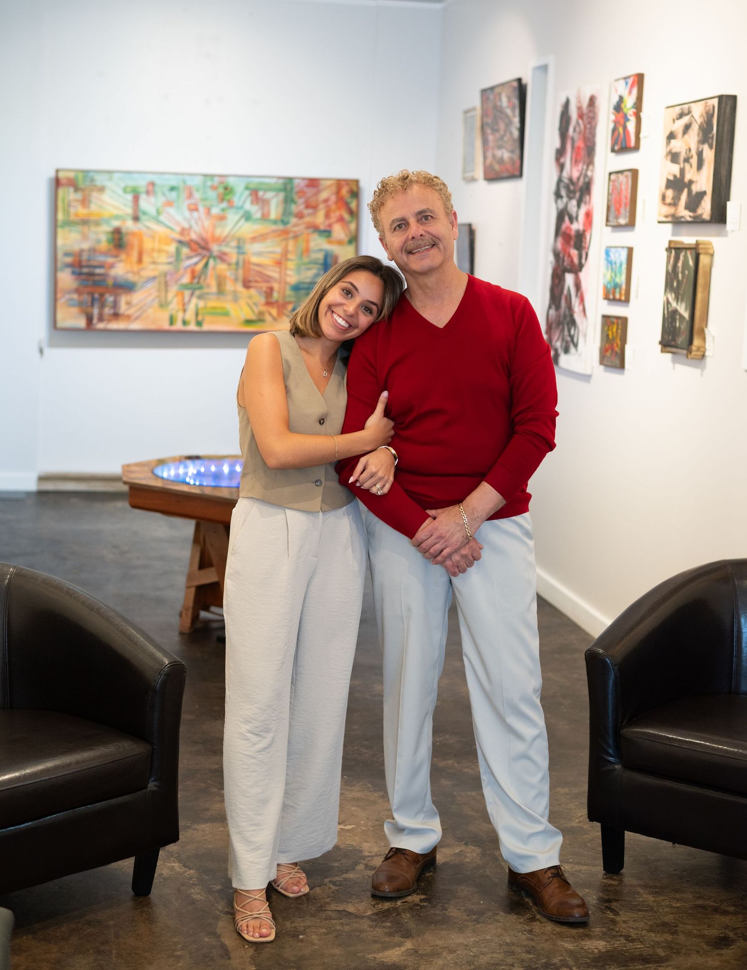 Smiling father and daughter embracing in a bright art gallery surrounded by colorful paintings on white walls.