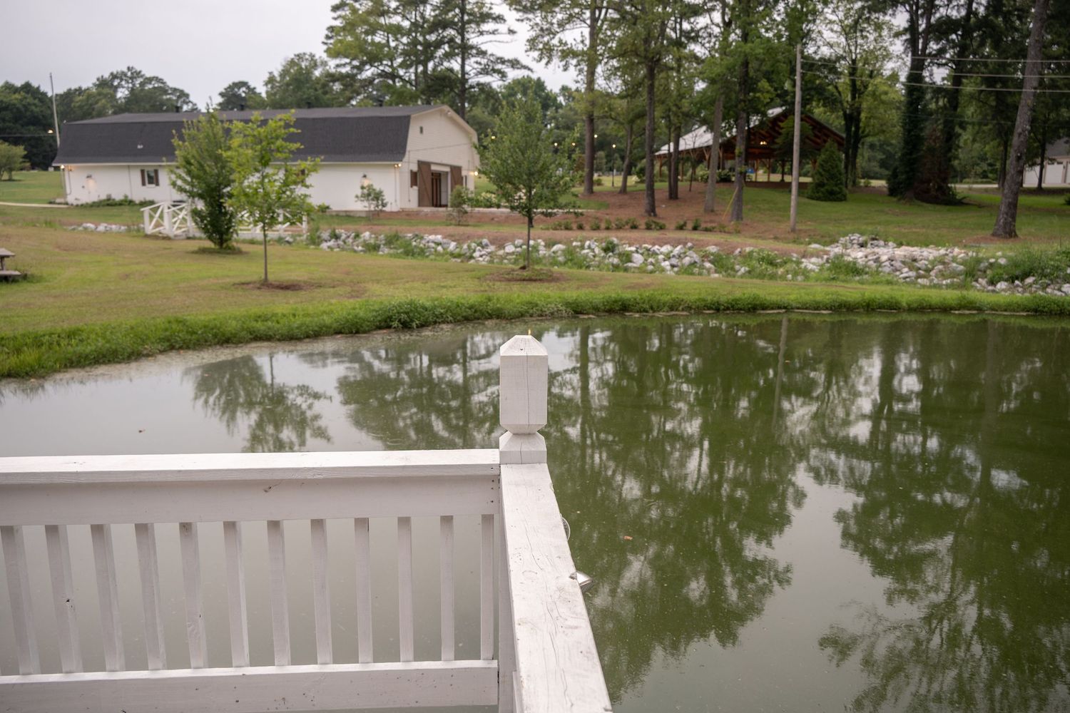 White railing overlooks a serene pond reflecting green trees, with a barn and pavilion on a lush rural property.