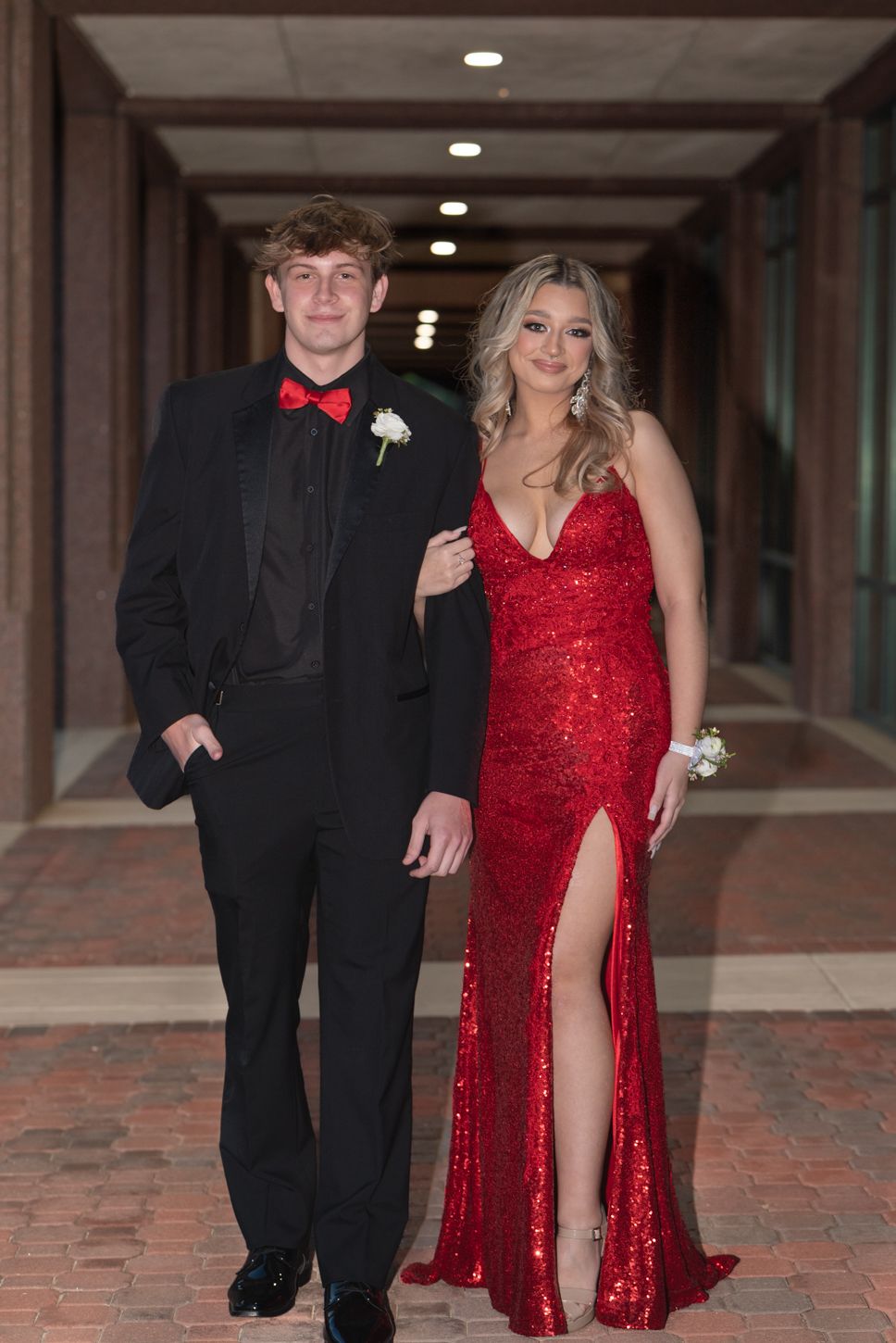 A young couple dressed in prom attire, man in black tuxedo with red bow tie, woman in red sequin gown, posing in hallway.