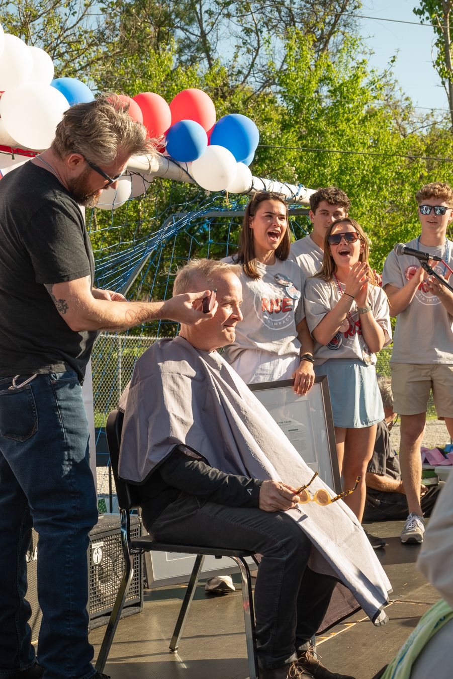 Man getting head shaved outdoors at charity event with cheering crowd and red, white, and blue balloons.