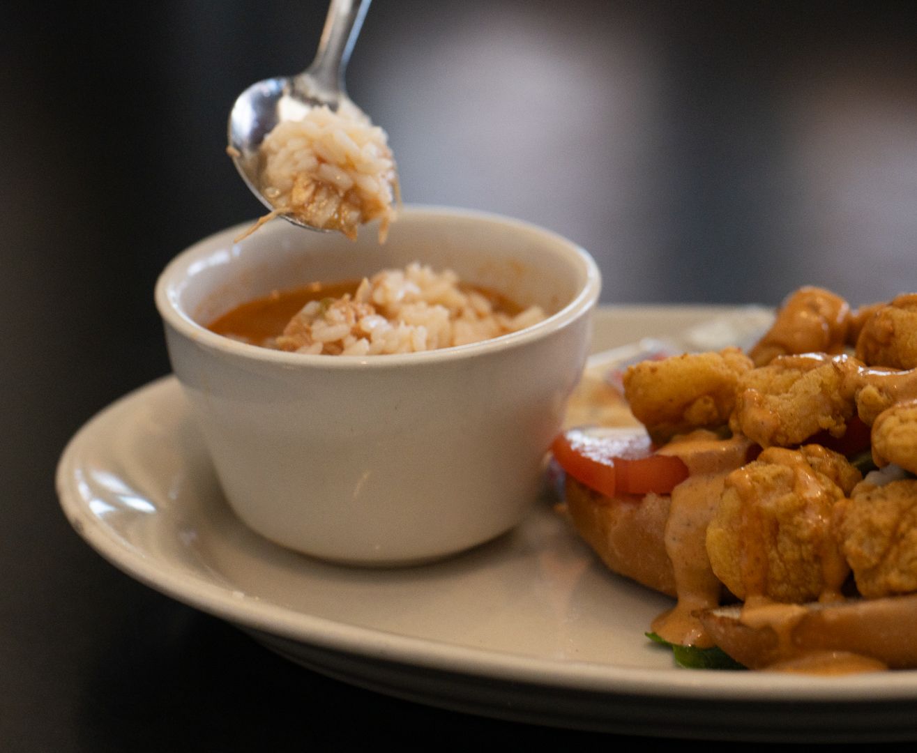 Spoon lifting rice over a bowl of sauce beside a shrimp po-boy.