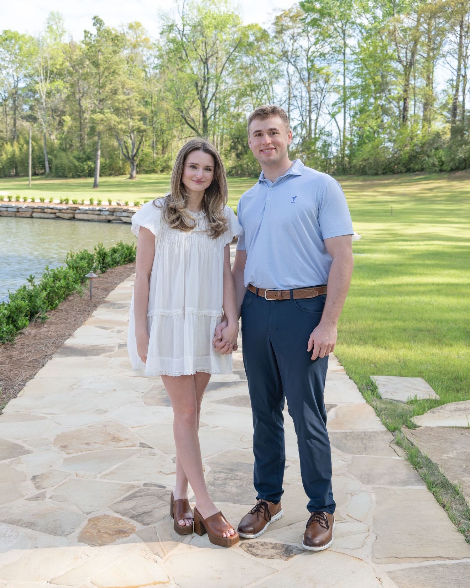 A young couple holds hands on a stone path by a lake, surrounded by lush greenery on a sunny day.