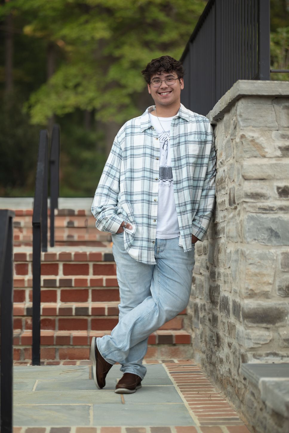 Smiling teenage boy in plaid shirt and jeans leaning against stone wall for senior portraits.