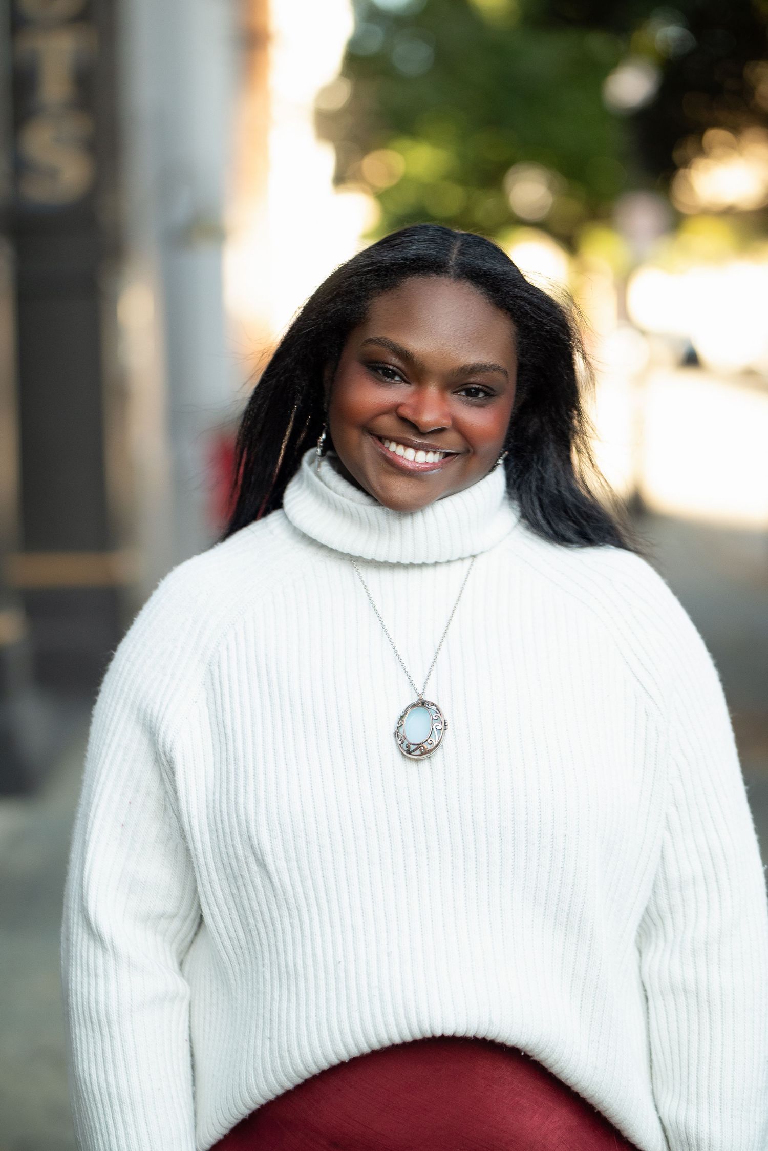 Smiling woman in white turtleneck sweater and red skirt posing on an urban street outdoors.